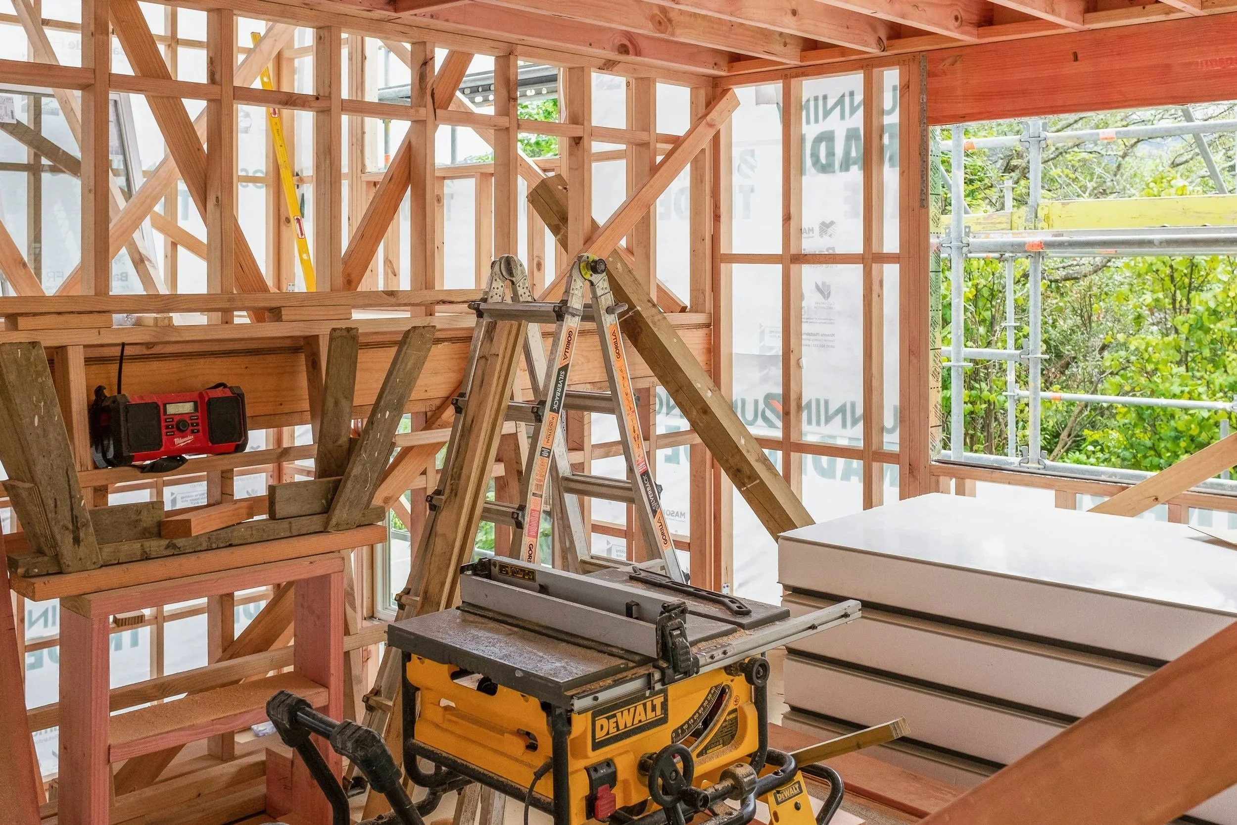 Interior view of a building under construction with exposed wooden framing, Bunnings site wrap, construction tools, and scaffolding outside visible through the windows.