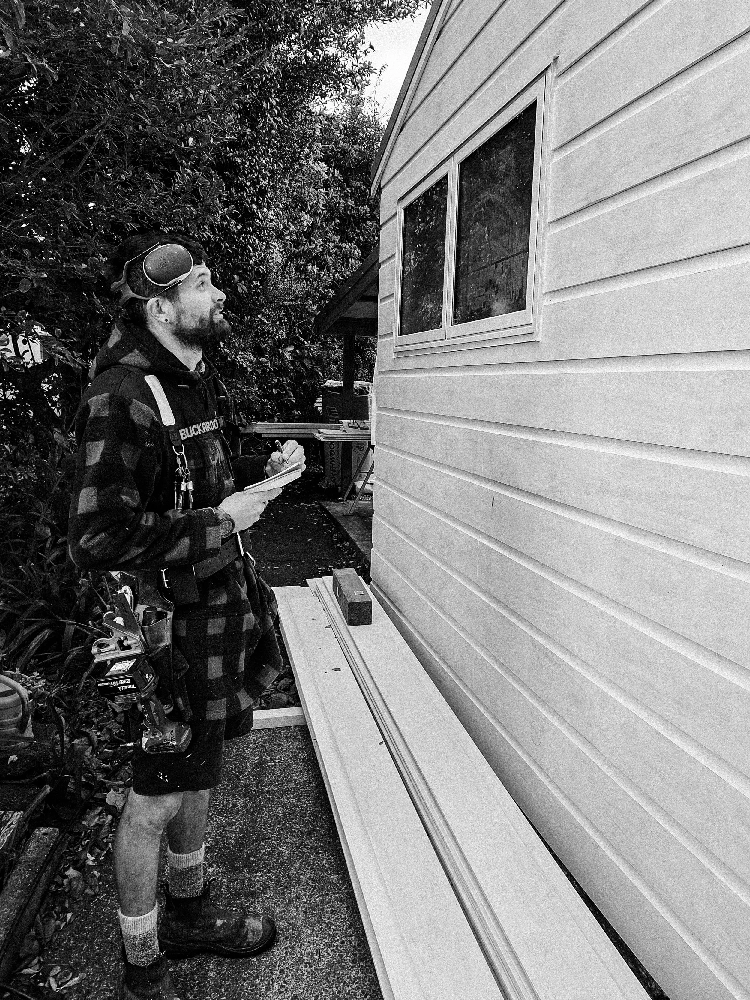 A man, wearing work clothes and a cap with goggles, is standing outdoors, holding a notepad and pen, looking at a house siding with several pieces of trim or siding material laid out on the ground beside him.