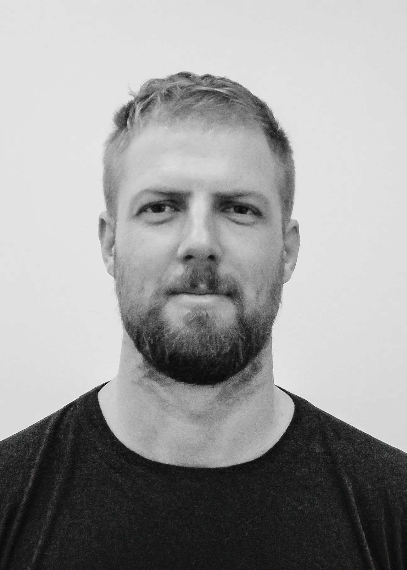 Black and white portrait of a man with a short beard and mustache, wearing a dark T-shirt, against a plain light background.