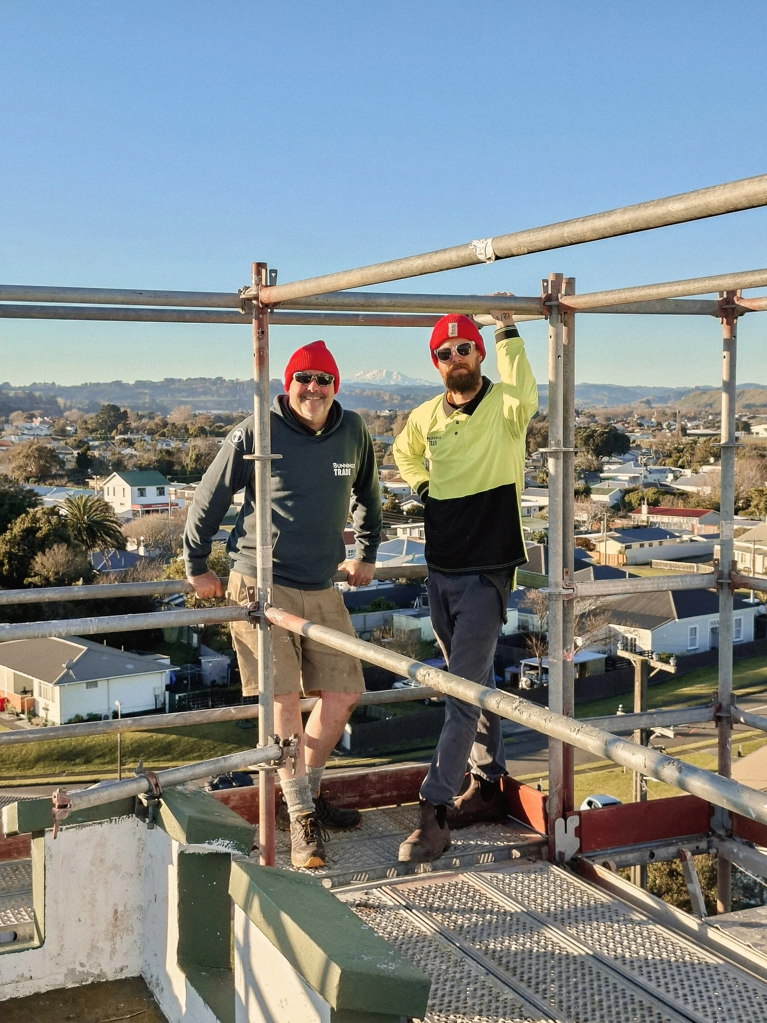 Two construction workers wearing red beanies and sunglasses standing on scaffolding with a cityscape and mountains in the background.
