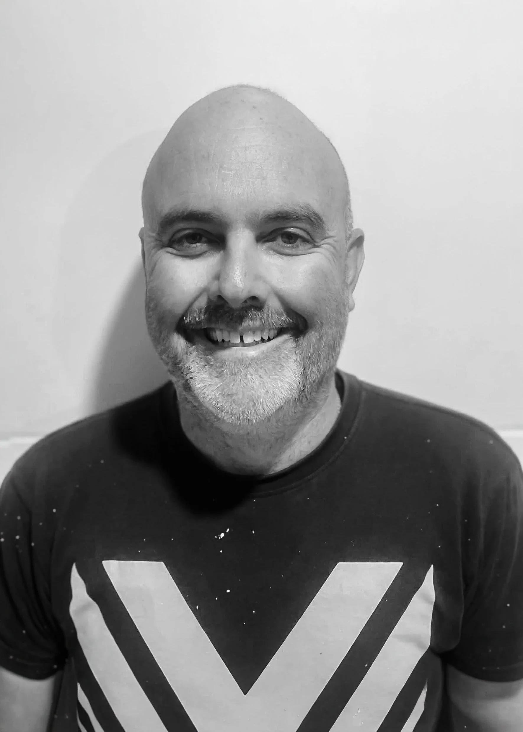 Black and white photo of a smiling, bald man with a beard and mustache, wearing a dark T-shirt, standing against a plain background.