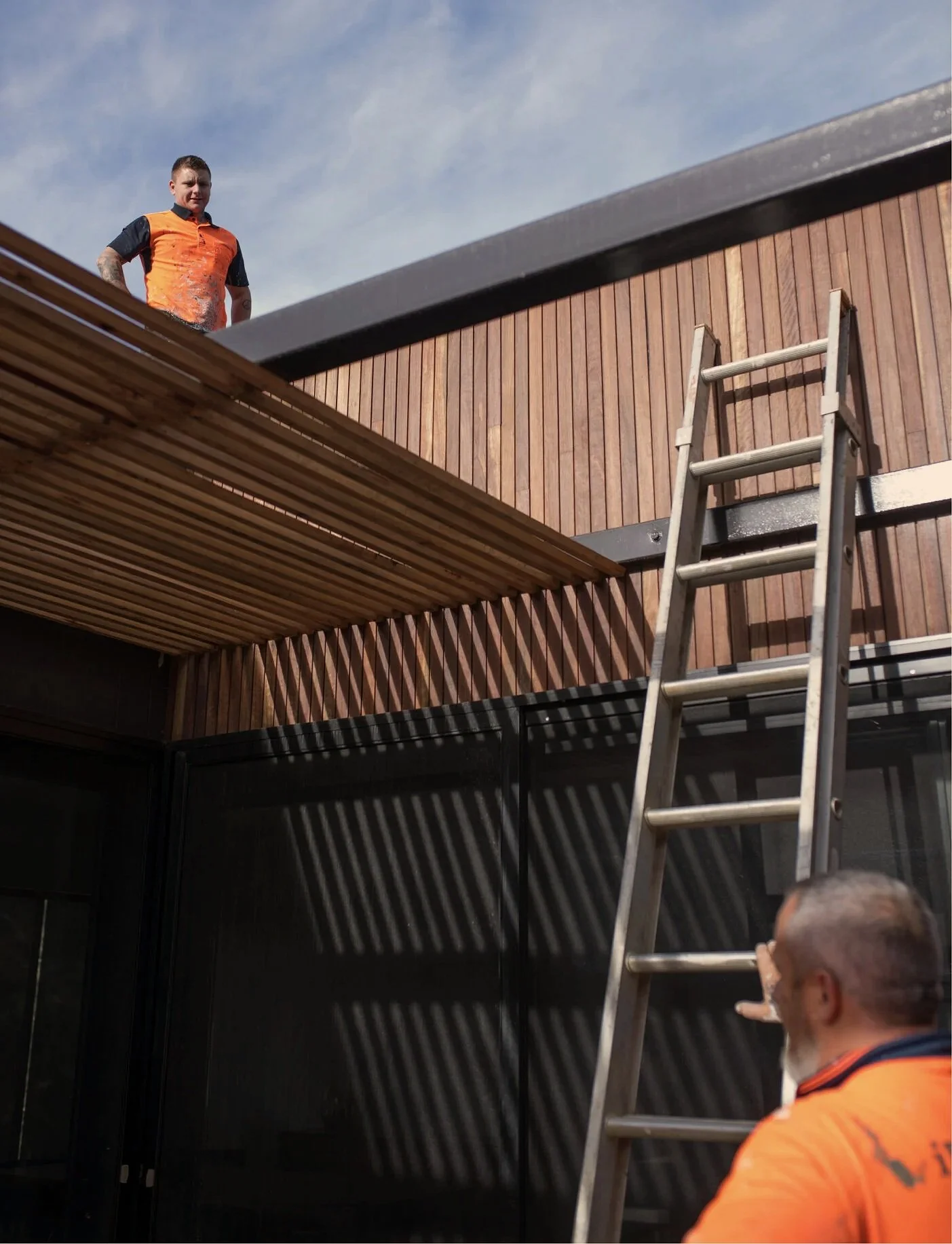 A man in an orange and black shirt stands on a roof under a partly cloudy sky, next to a wooden building with vertical planks, a ladder leaning against it, and a black wall below.
