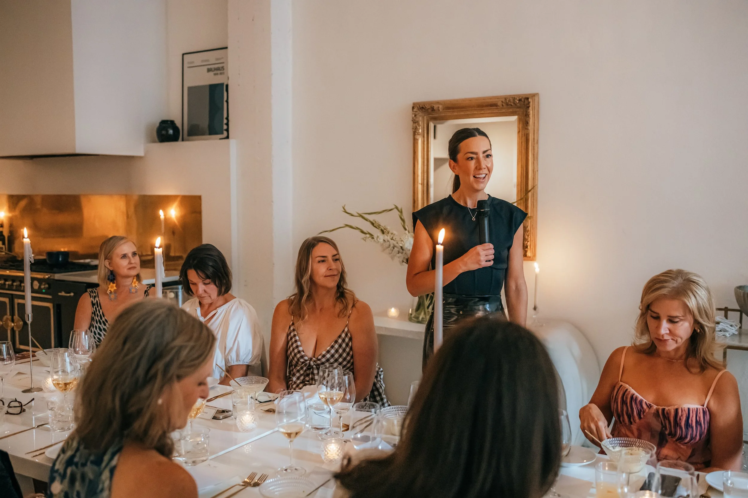 Megan Gonyo in a black dress is speaking into a microphone at a dinner event. Several women are seated around a table with candles and wine glasses, listening to her. The room has white walls, a gold-framed mirror, and decorative items.