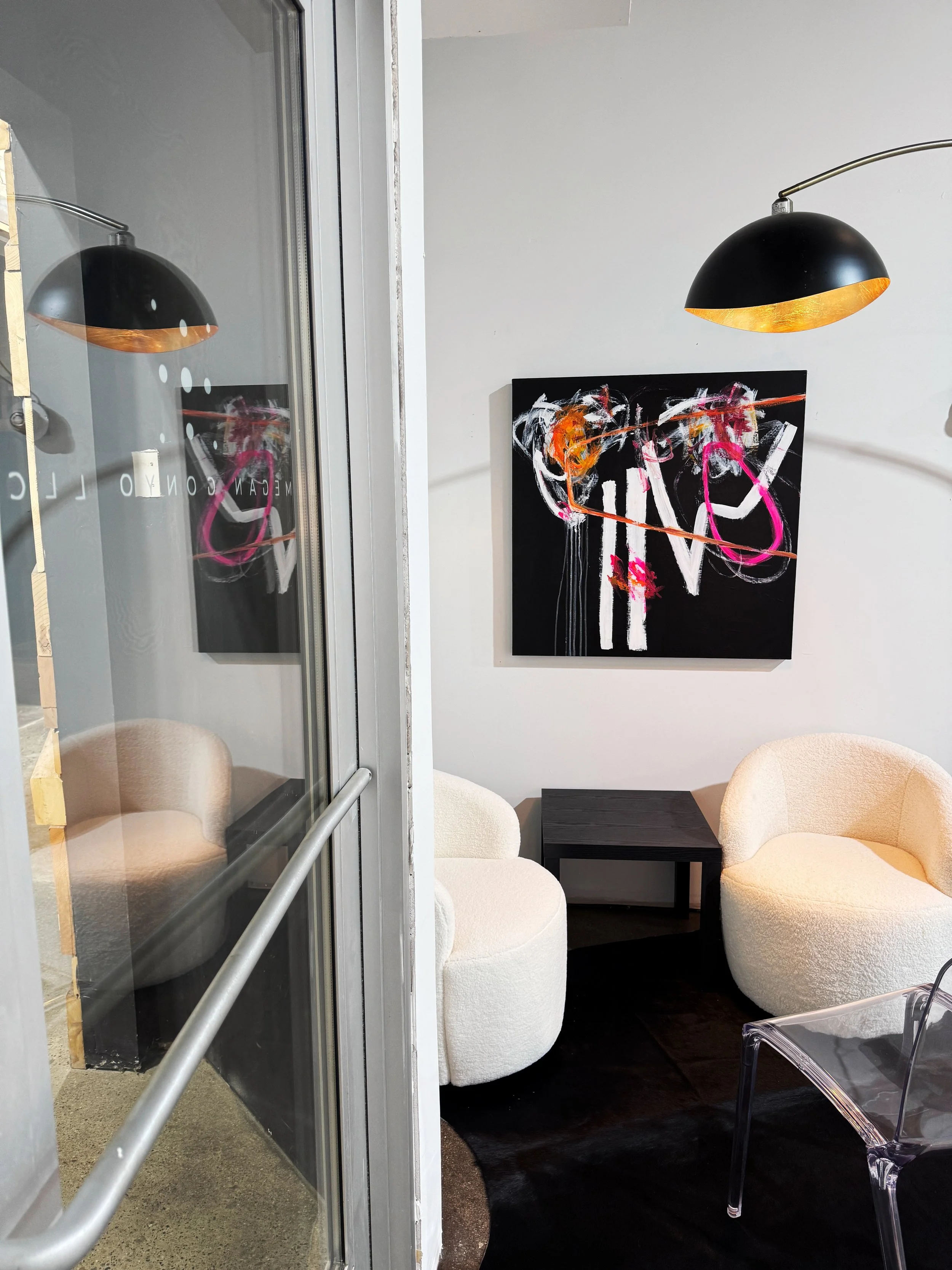 A cozy corner in Megan Gonyo's art studio with two plush, white, rounded armchairs and a black side table, decorated with vibrant abstract art on the wall, adjacent to a glass wall reflecting a black lamp with a gold interior.