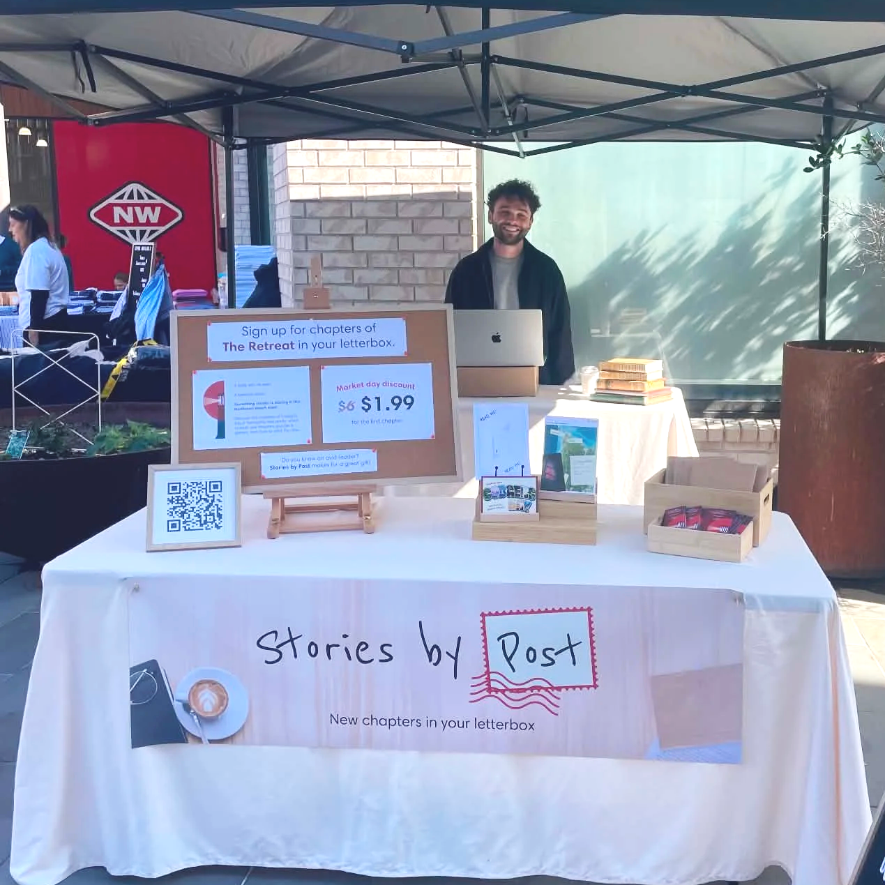 A man standing behind a table at an outdoor event, smiling. The table has a sign promoting new chapters in your letterbox, and features books, brochures, and a QR code. There is a can of drink on the table and a laptop in front of the man. Other tables and people are visible in the background under a canopy.