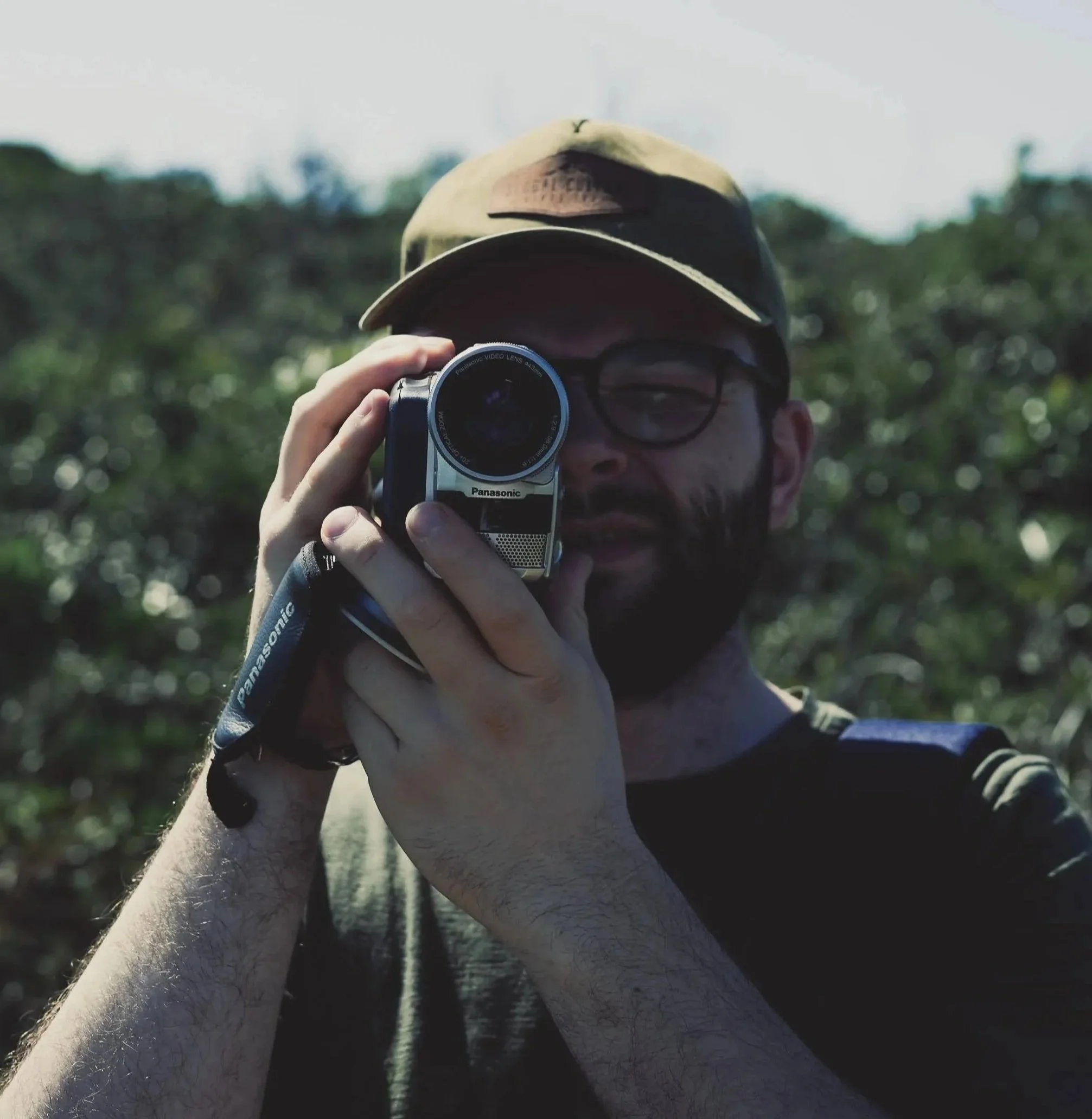 Person holding a camera taking a selfie, wearing a baseball cap, glasses, in an outdoor setting with trees and sky background.