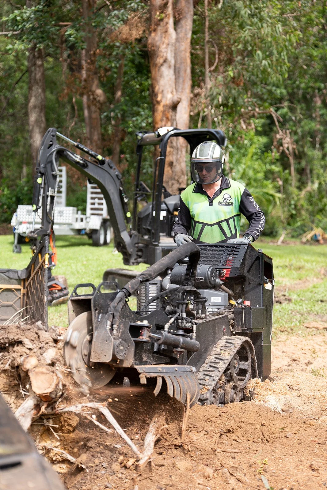 An arborist wearing a helmet and safety gear operates a small stump grinder in a wooded outdoor area, with trees and a vehicle in the background.