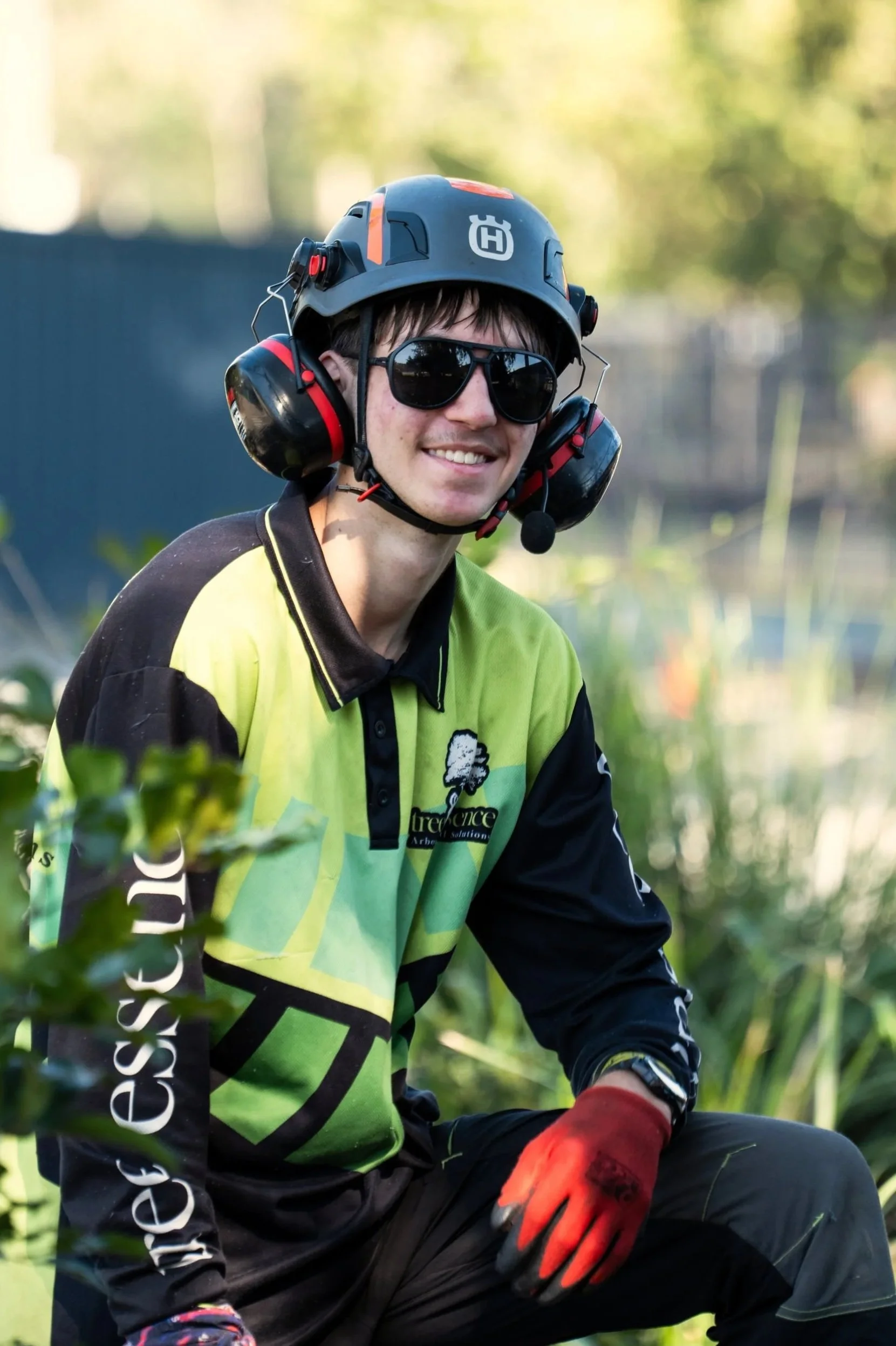 A man wearing a gray helmet with a red stripe, black sunglasses, a green and black long-sleeved shirt, and red gloves sitting outdoors amidst green foliage.