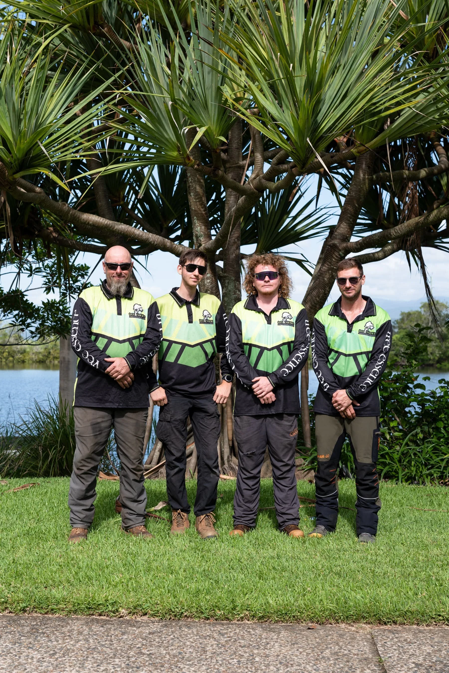 Four men wearing matching black and green shirts with logos stand outdoors in front of a large tree with long green leaves, near water and green grass.