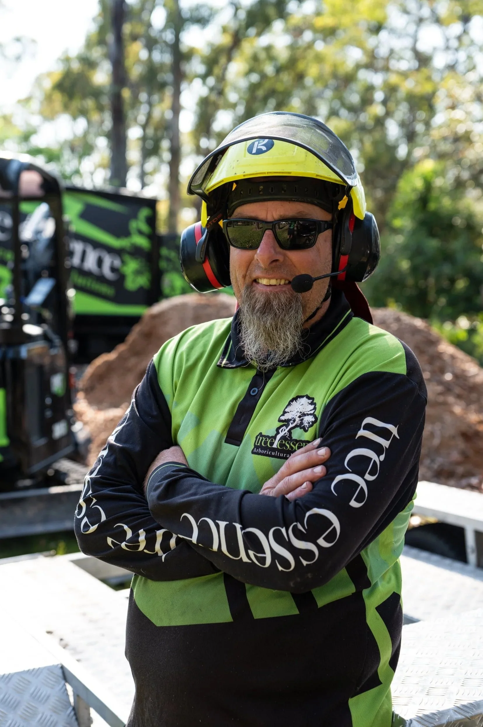 A man wearing safety gear, including a helmet with a visor, sunglasses, and a headset with microphone, stands outdoors. He is dressed in a green and black long-sleeved shirt with a logo on the chest, with crossed arms, in front of a background of trees and some machinery.