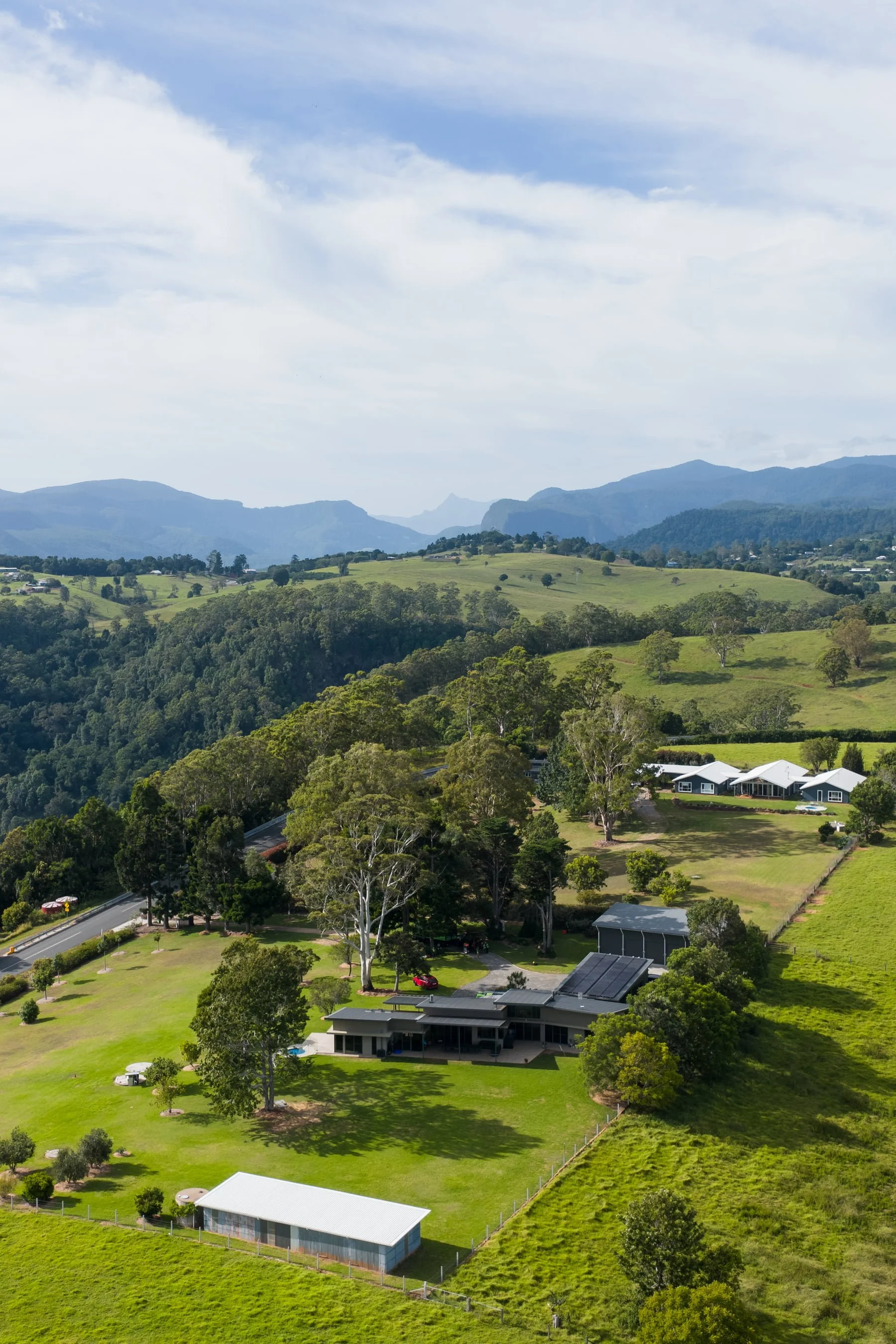 Scenic view of rolling green hills, scattered trees, houses, and mountains in the background under a partly cloudy sky.