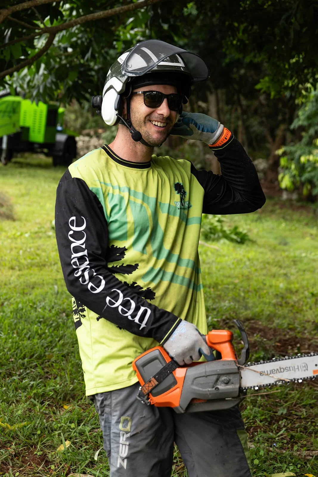 Smiling arborist wearing safety goggles, helmet, and gloves, holding a chainsaw outdoors in a grassy, wooded area.