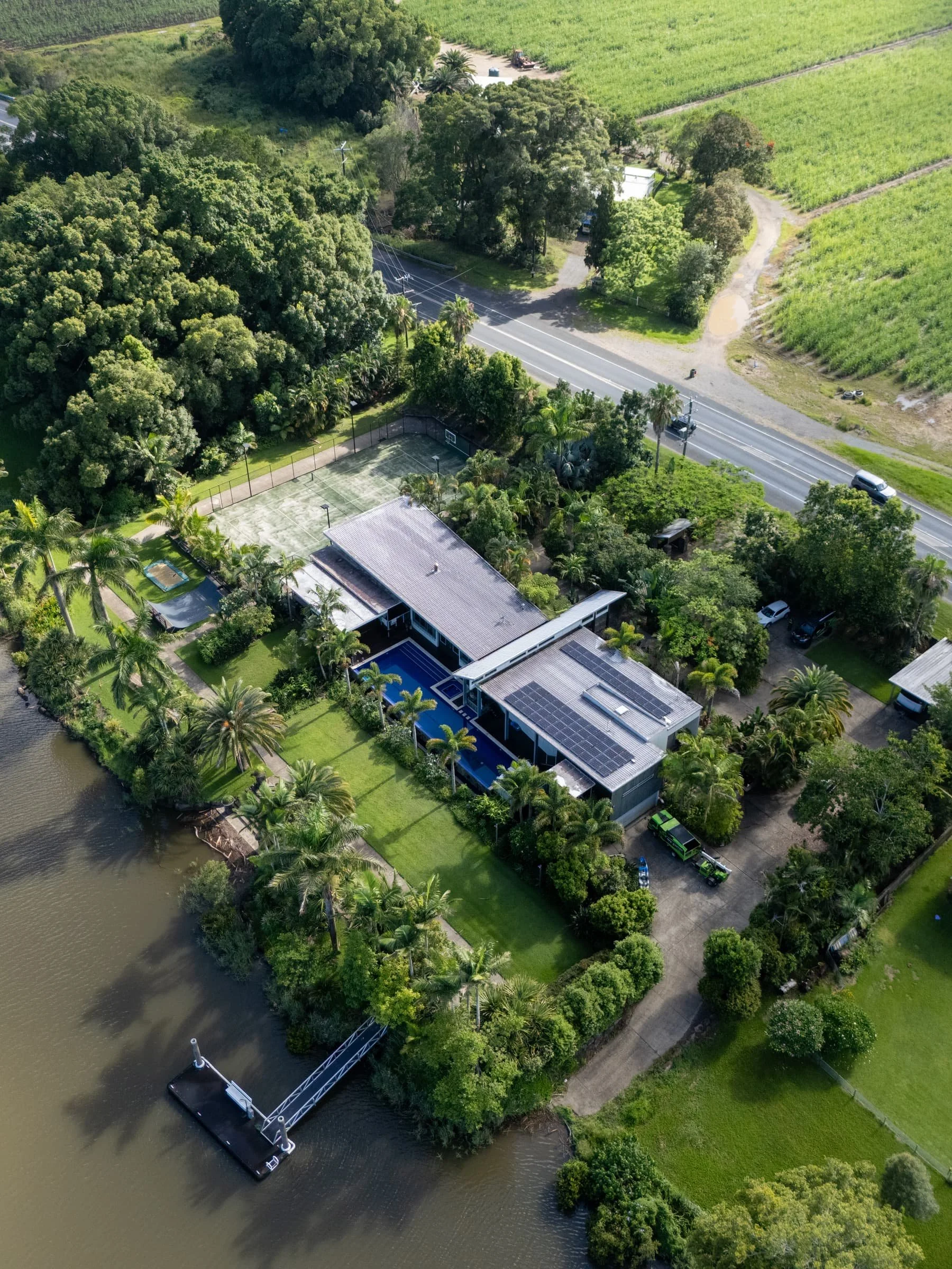 Aerial view of a house with a swimming pool, tennis court, boat dock, and lush green palm trees along a river, with roads and farmland nearby.