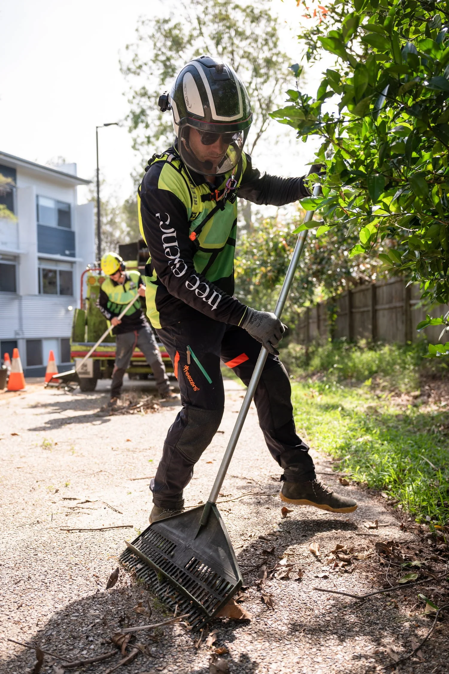 Two workers in safety gear, including helmets and high-visibility shirts, are working outdoors on a footpath. One worker is using a rake to clear debris and leaves from the ground, while the other is in the background adding debris to a wood chipper.