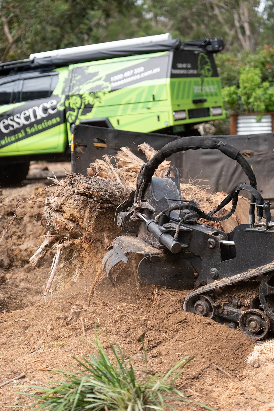 Close-up of a stump grinder around a tree root on dirt, with a green and black vehicle labeled 'Tree Essence' in the background.