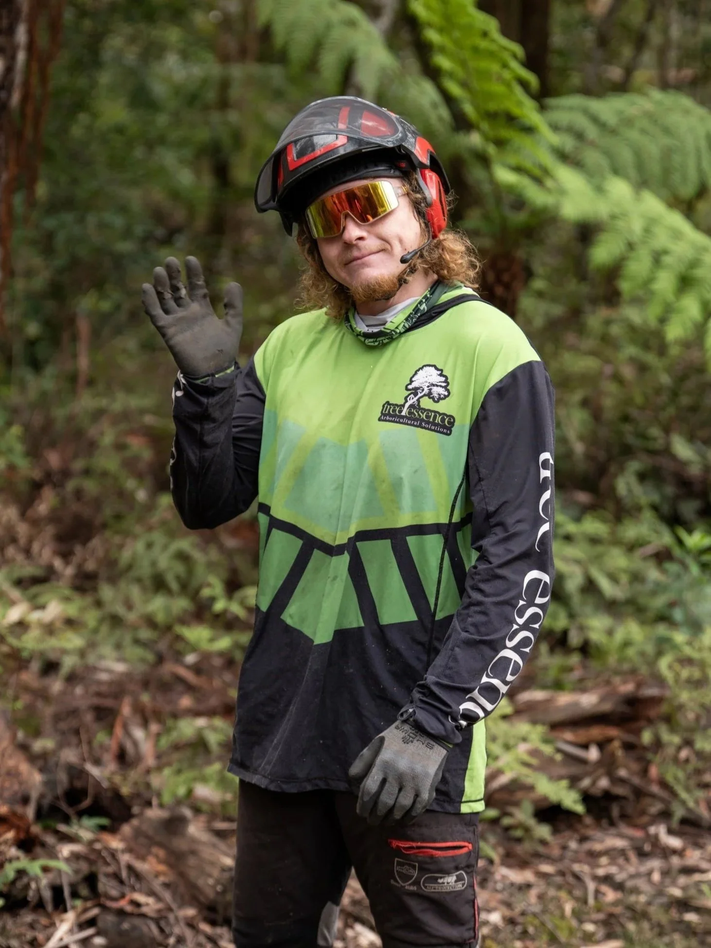 Arborist in forest wearing helmet, sunglasses, gloves, and green and black long-sleeved shirt, waving.