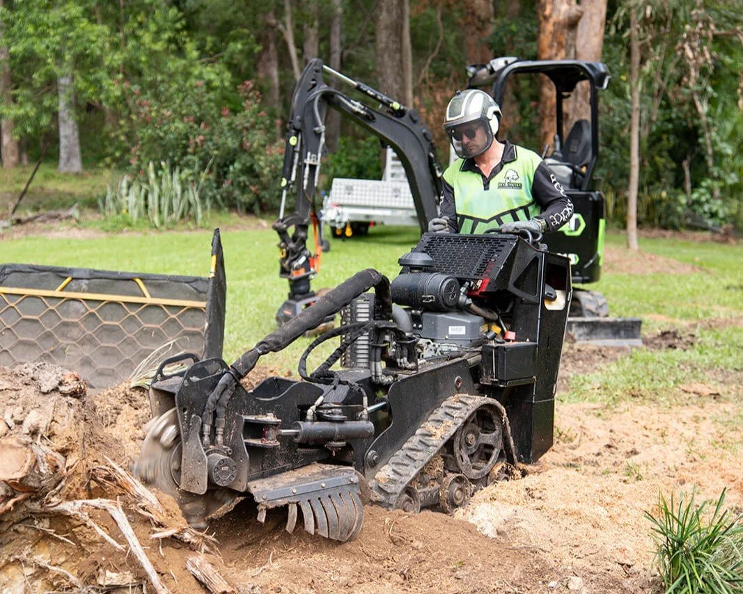 An arborist wearing a safety helmet and sunglasses operates a stump grinder in a grassy area with trees in the background. The stump grinder is digging into the stump in the ground.