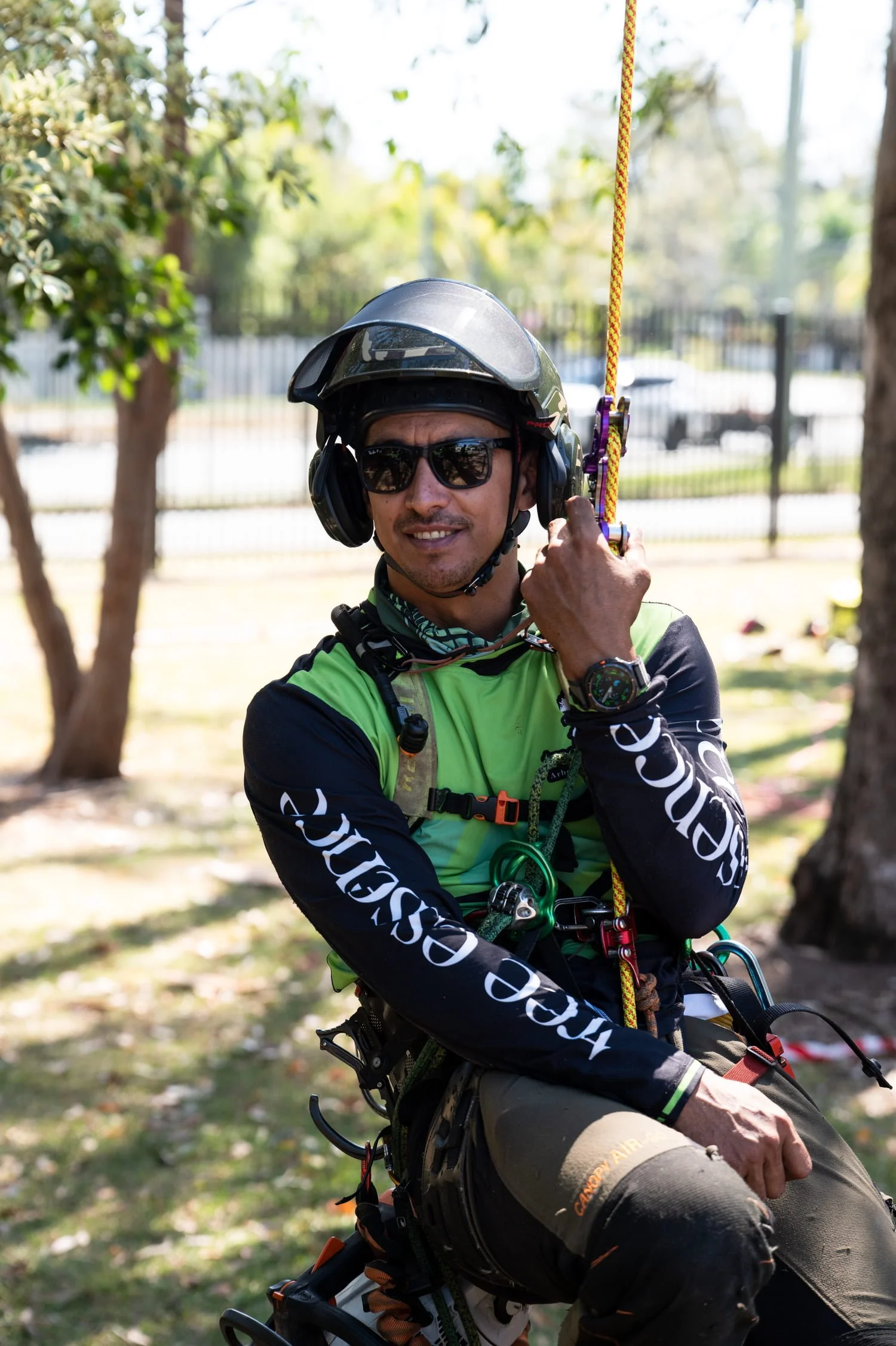 Arborist in a helmet and sunglasses sitting from arborist climbing ropes outdoors, holding a climbing rope, with trees and a fence in the background.