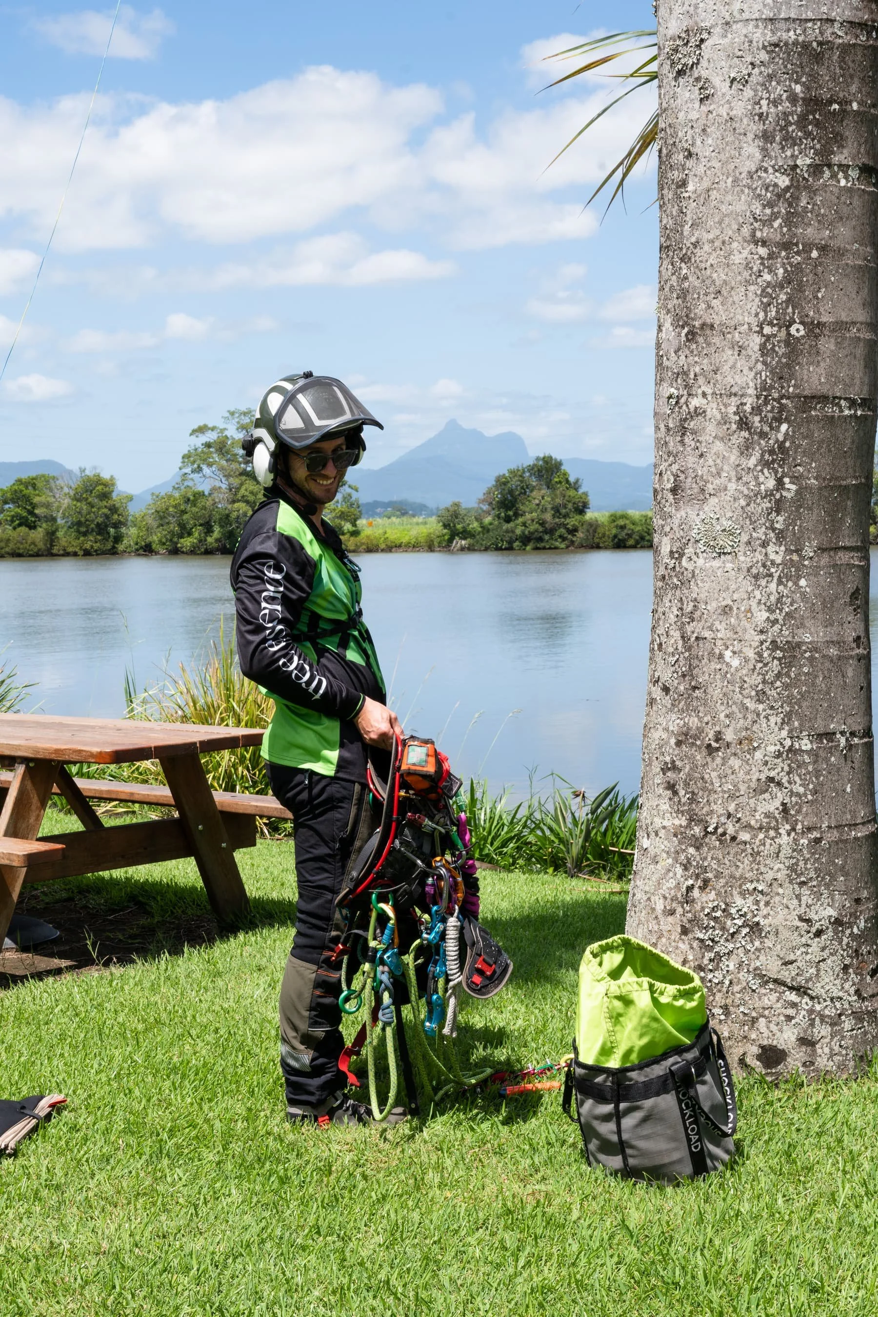 A man wearing a green and black climbing jacket, helmet, sunglasses, and climbing gear stands outdoors by a tree near a river, with mountains and blue sky in the background. He is smiling and preparing climbing equipment, with a backpack and climbing accessories on the grass nearby.