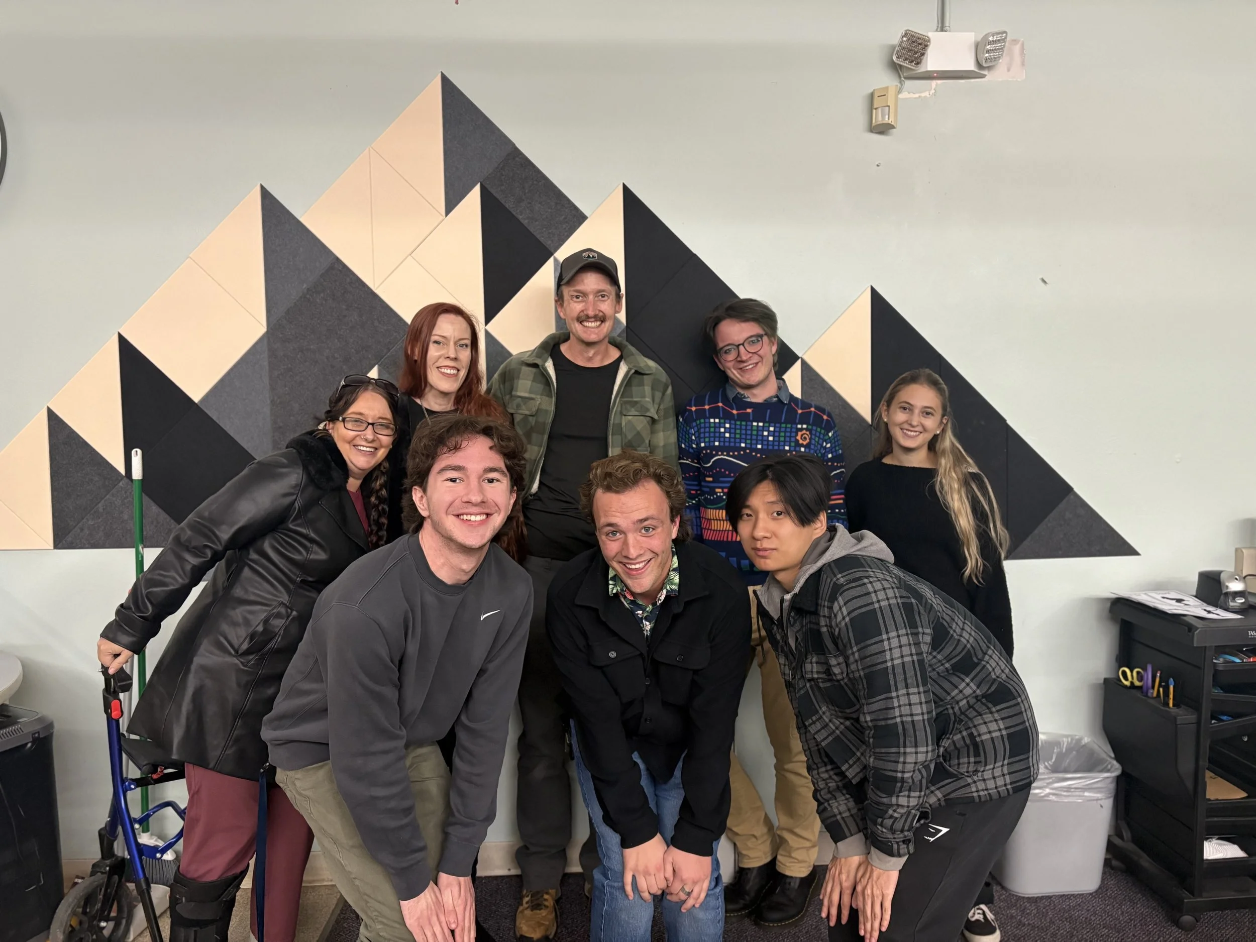A group of eight people smiling and posing together in a room with a geometric dark and beige wall design.