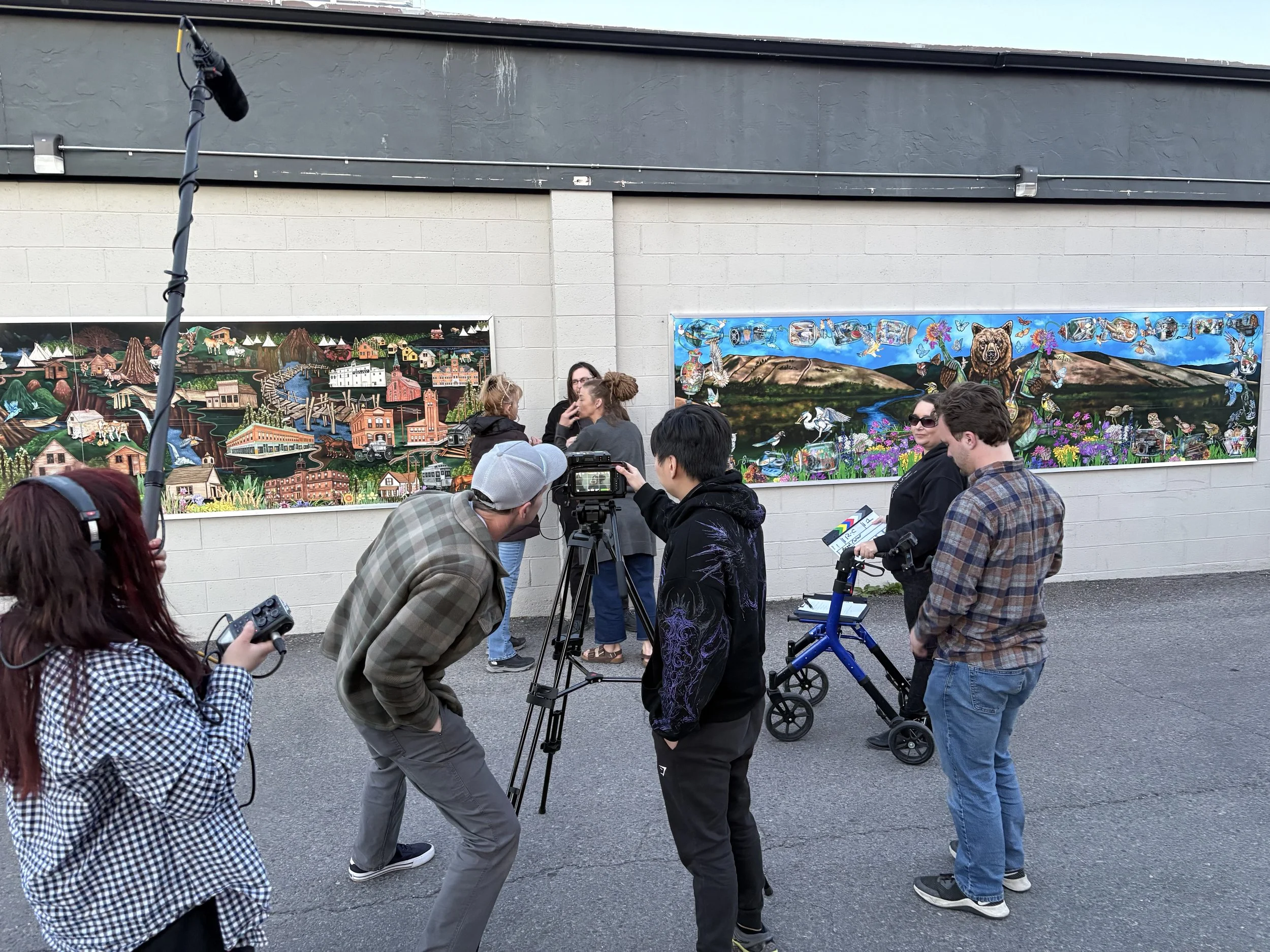 A group of people gathered outdoors, many holding cameras or phones, surrounding a mural painted on a wall. The mural depicts a colorful landscape with buildings, mountains, trees, animals, and butterflies, with some people in conversation in front of it.