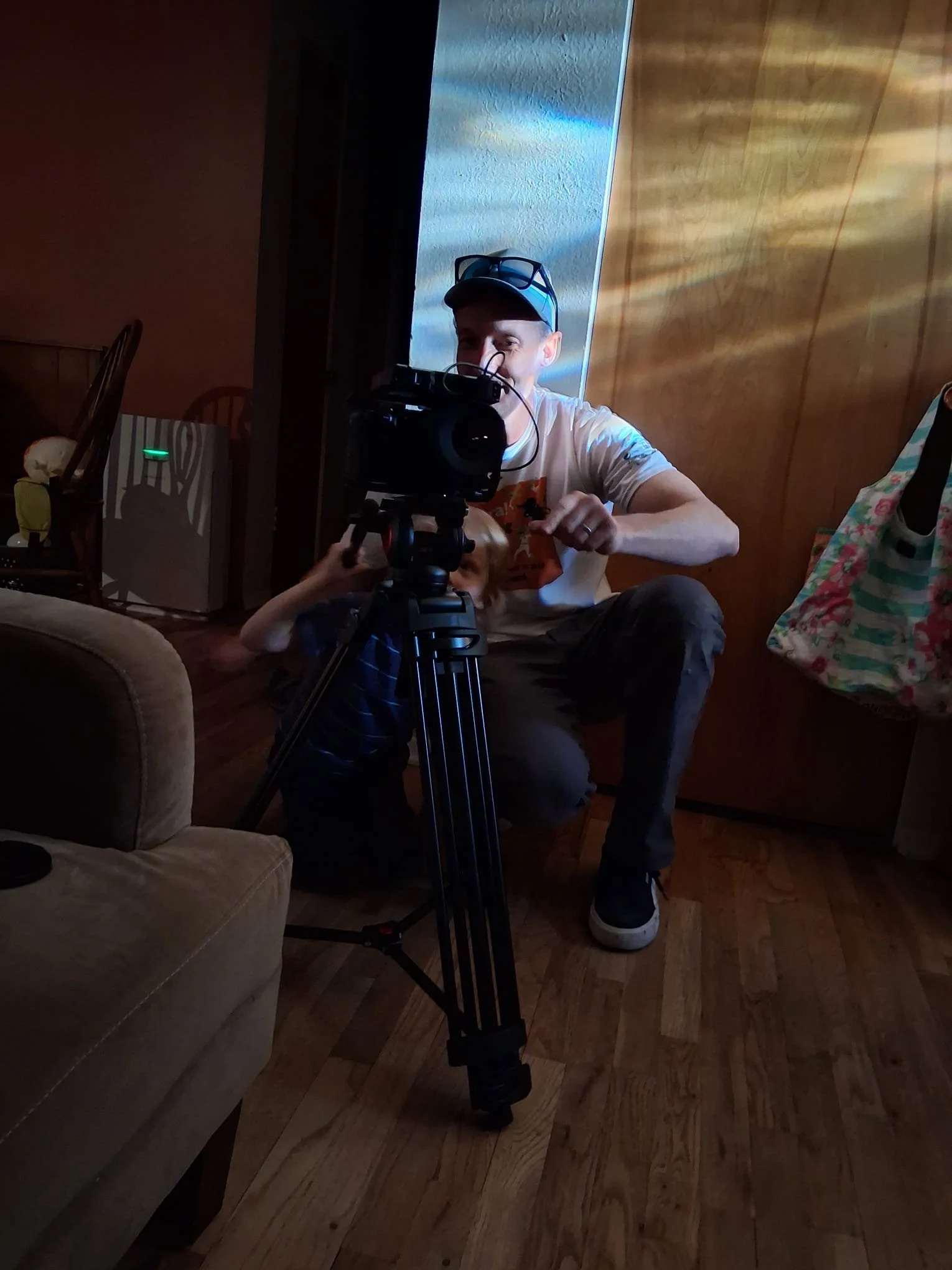 A person wearing a cap, glasses, and a beige T-shirt is adjusting camera equipment while sitting on the floor in a room with wooden panel walls and a hardwood floor. There is a beige couch, a chair, an air purifier, and a colorful bag in the background.
