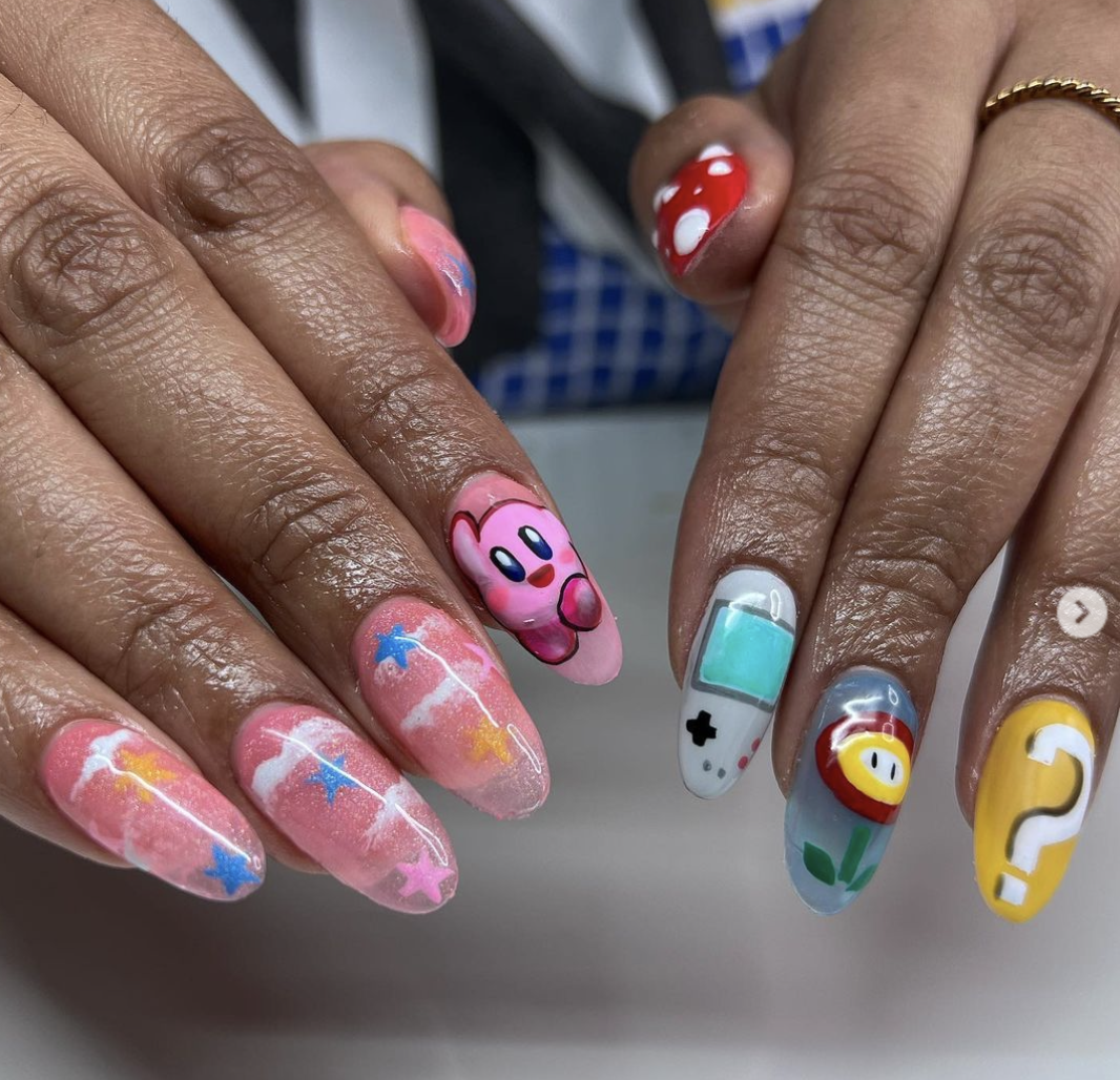 Nail art with cartoon characters and playful designs, including Kirby, a smiley face flower, a question mark, a handheld gaming device, a milk bottle, and a starry sky theme.