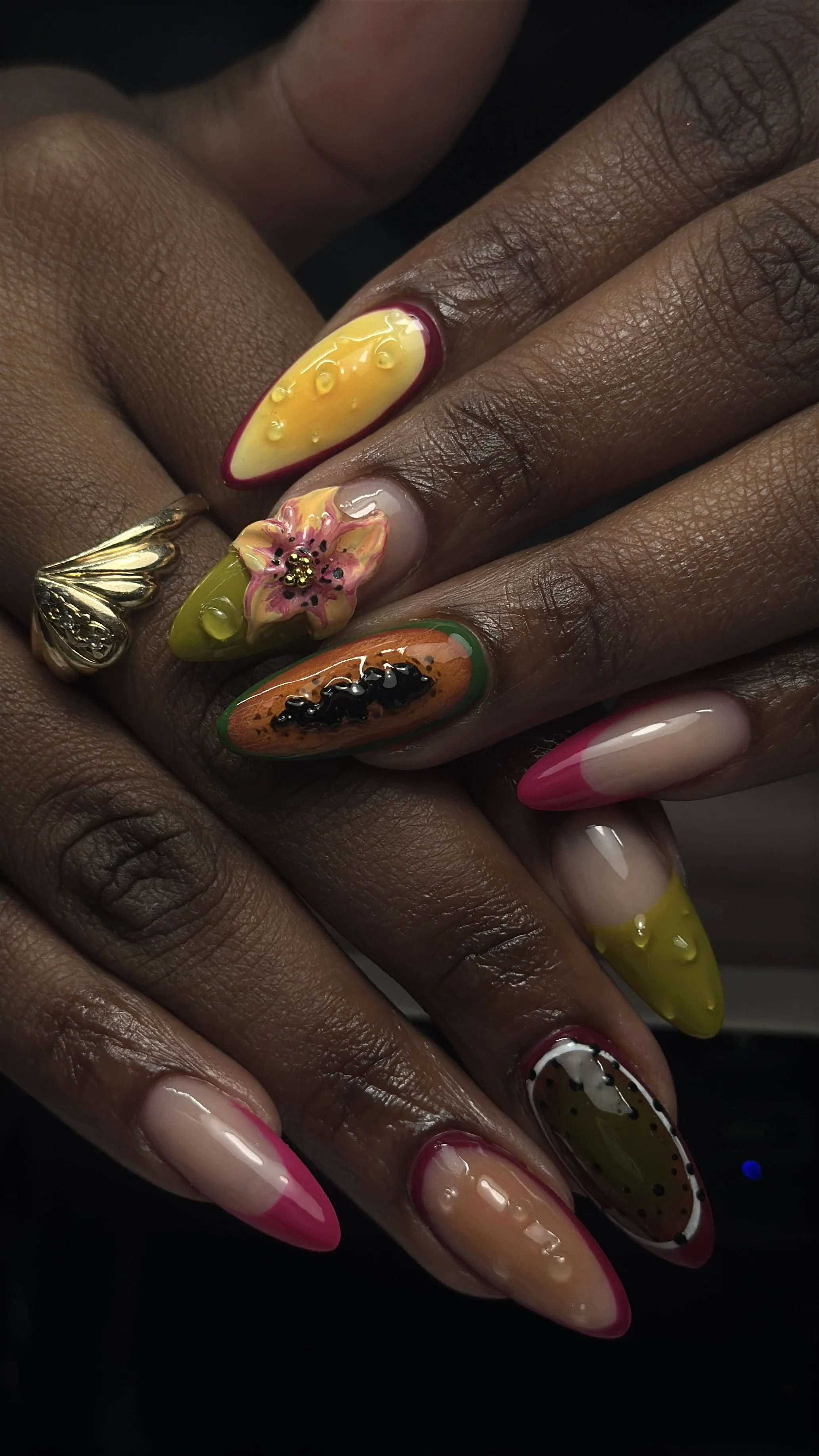 Close-up of hands with colorful, fruit-themed nail art, including designs resembling a banana, strawberry, pineapple, kiwi, and orange, with detailed embellishments.