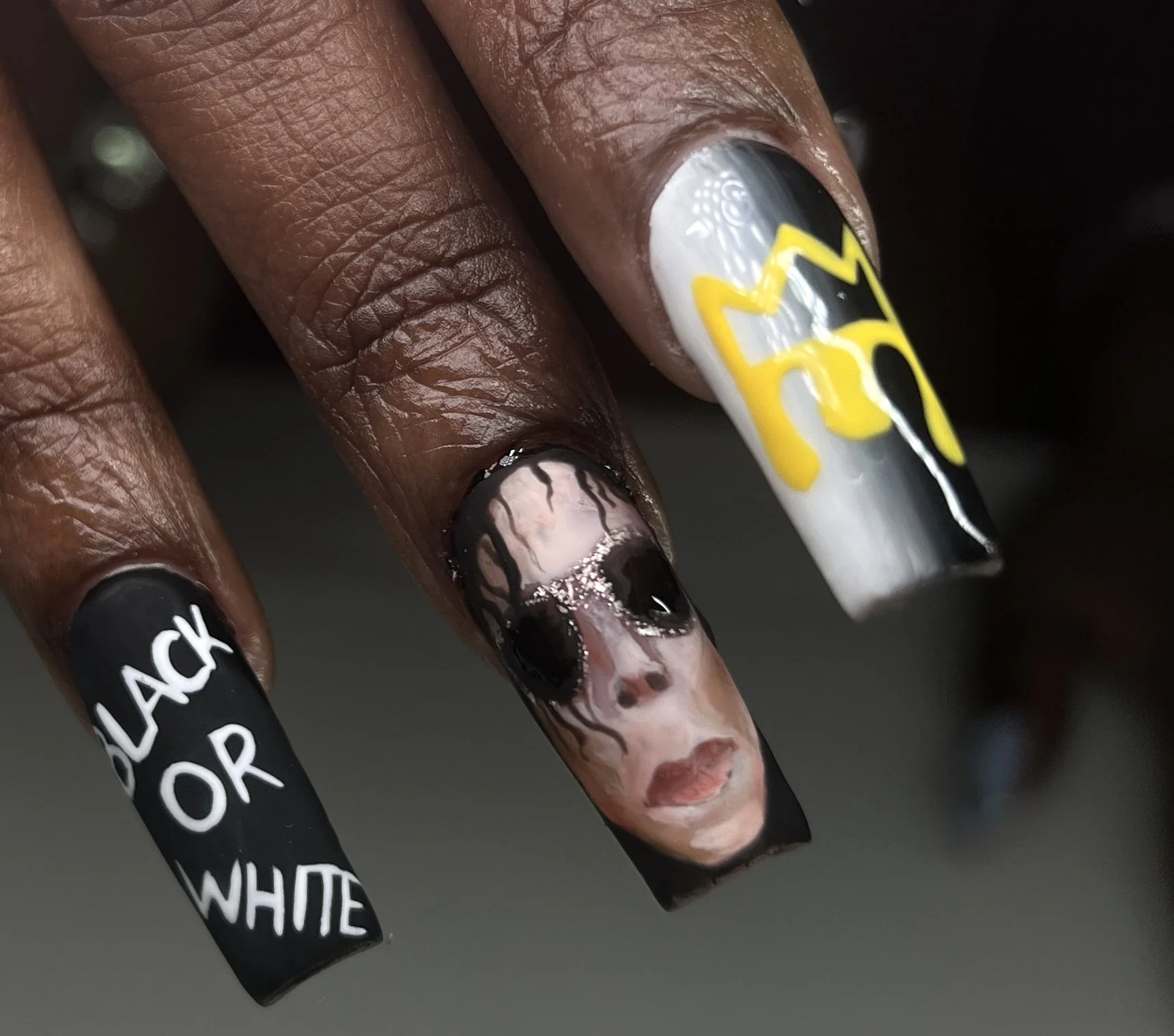 Close-up of three long, decorated fingernails with black, white, yellow, and pink nail polish and artwork, including the words 'Black or White' and a painted portrait of a Michael Jackson's face