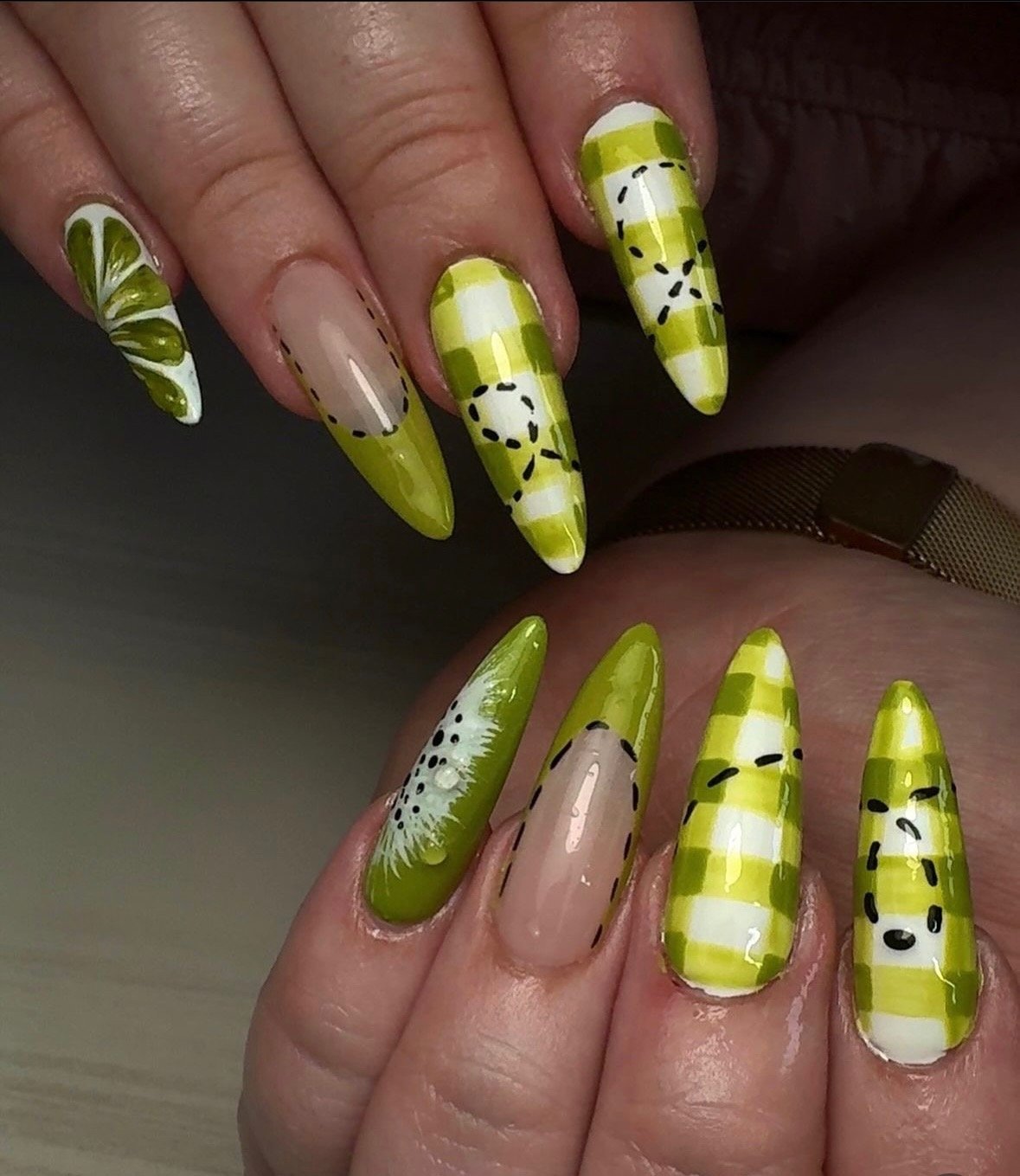 Close-up of hands with long, stiletto-shaped nails painted in a yellow-green plaid pattern with black dashed lines and white accents, some nails feature floral or leaf designs.