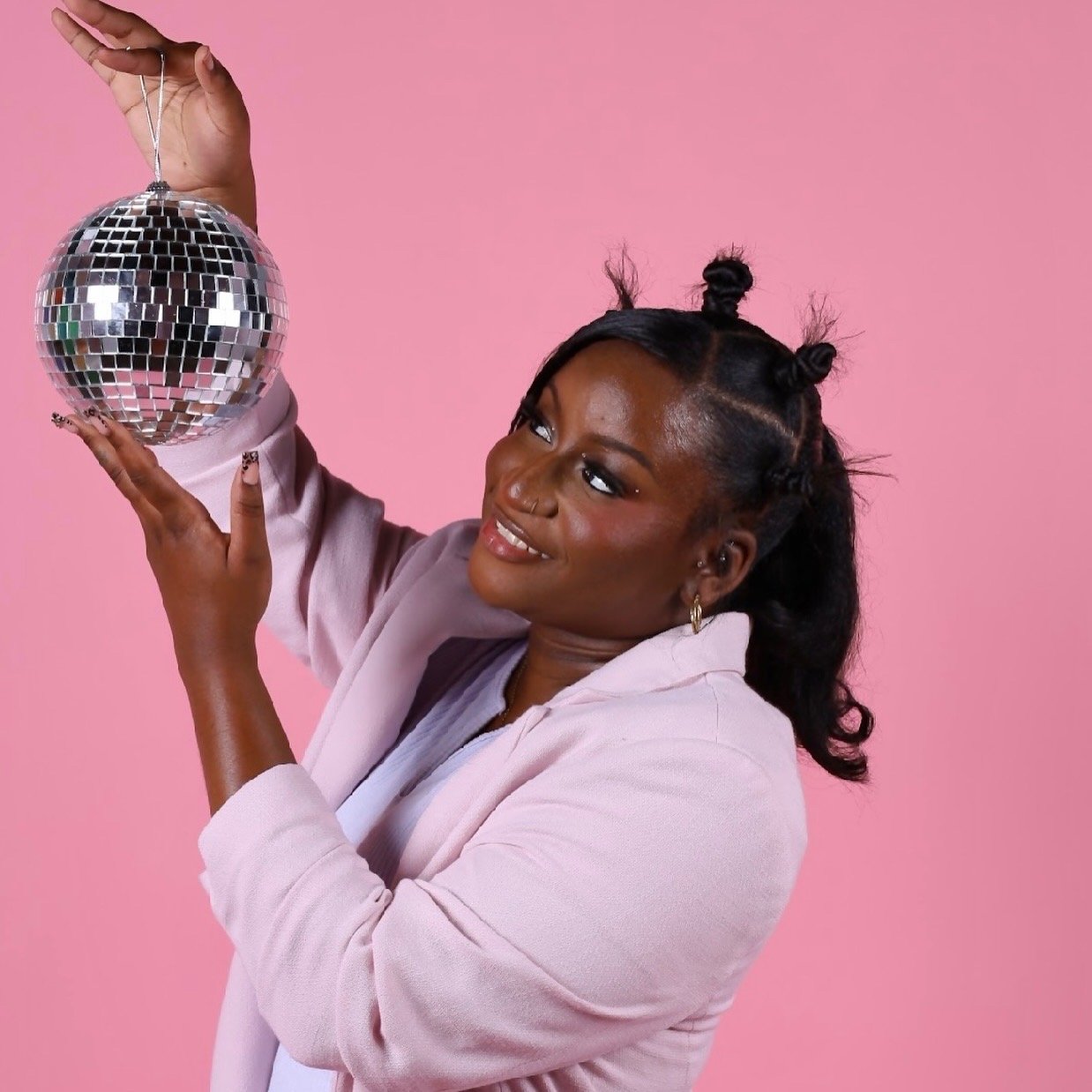 A woman with dark hair styled in small buns, wearing a pink blazer, holding a disco ball ornament against a pink background, smiling and looking at it.