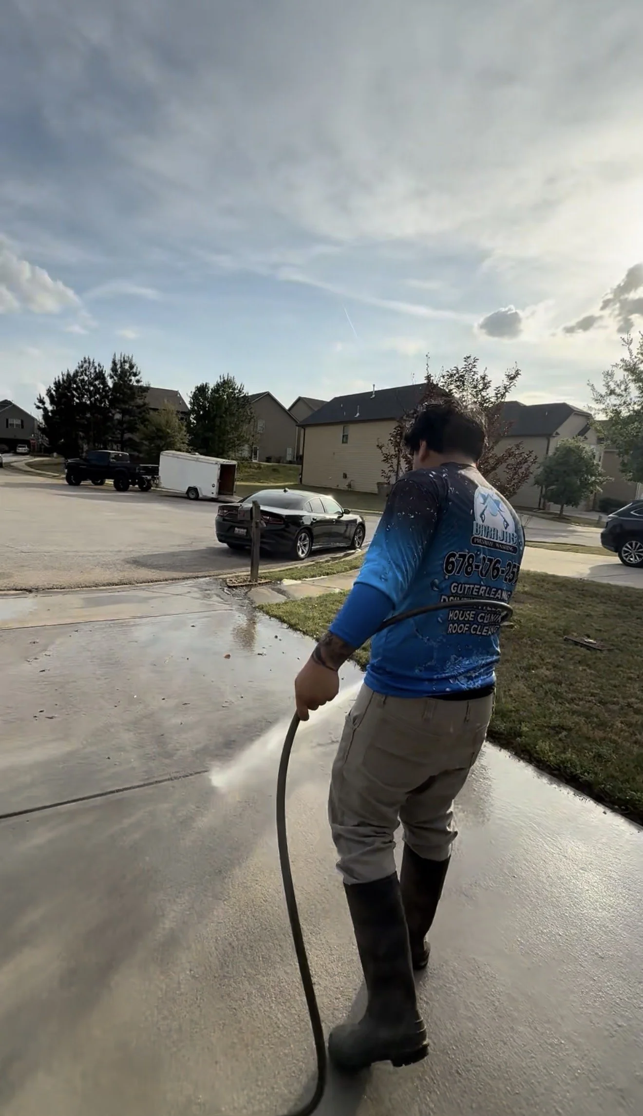 Worker in a blue uniform pressures washing a sidewalk in a residential neighborhood on a cloudy day.