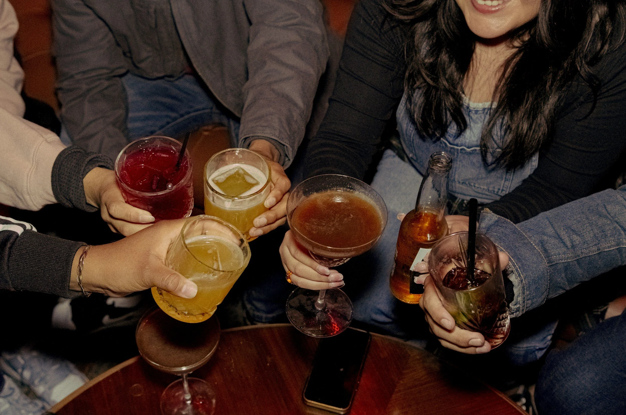 A group of friends raising various colorful drinks in a toast at a social gathering.