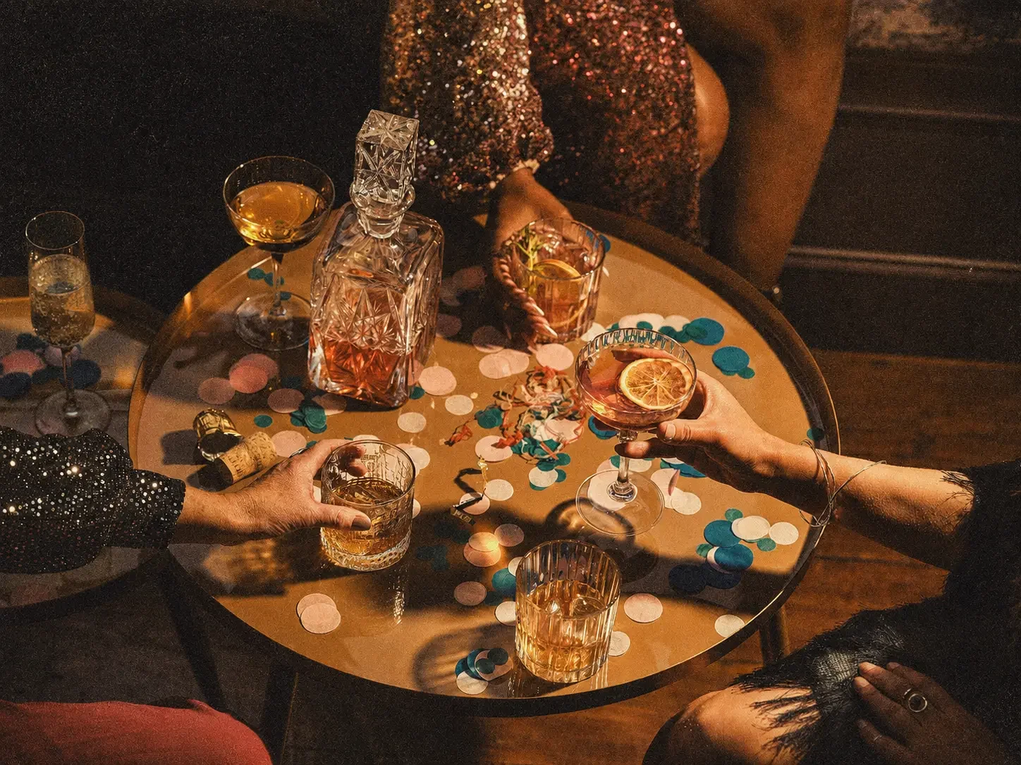People celebrating with drinks at a party, with confetti on a round table, including various glasses and bottles, some with lemon slices, and two hands visible, one holding a drink.