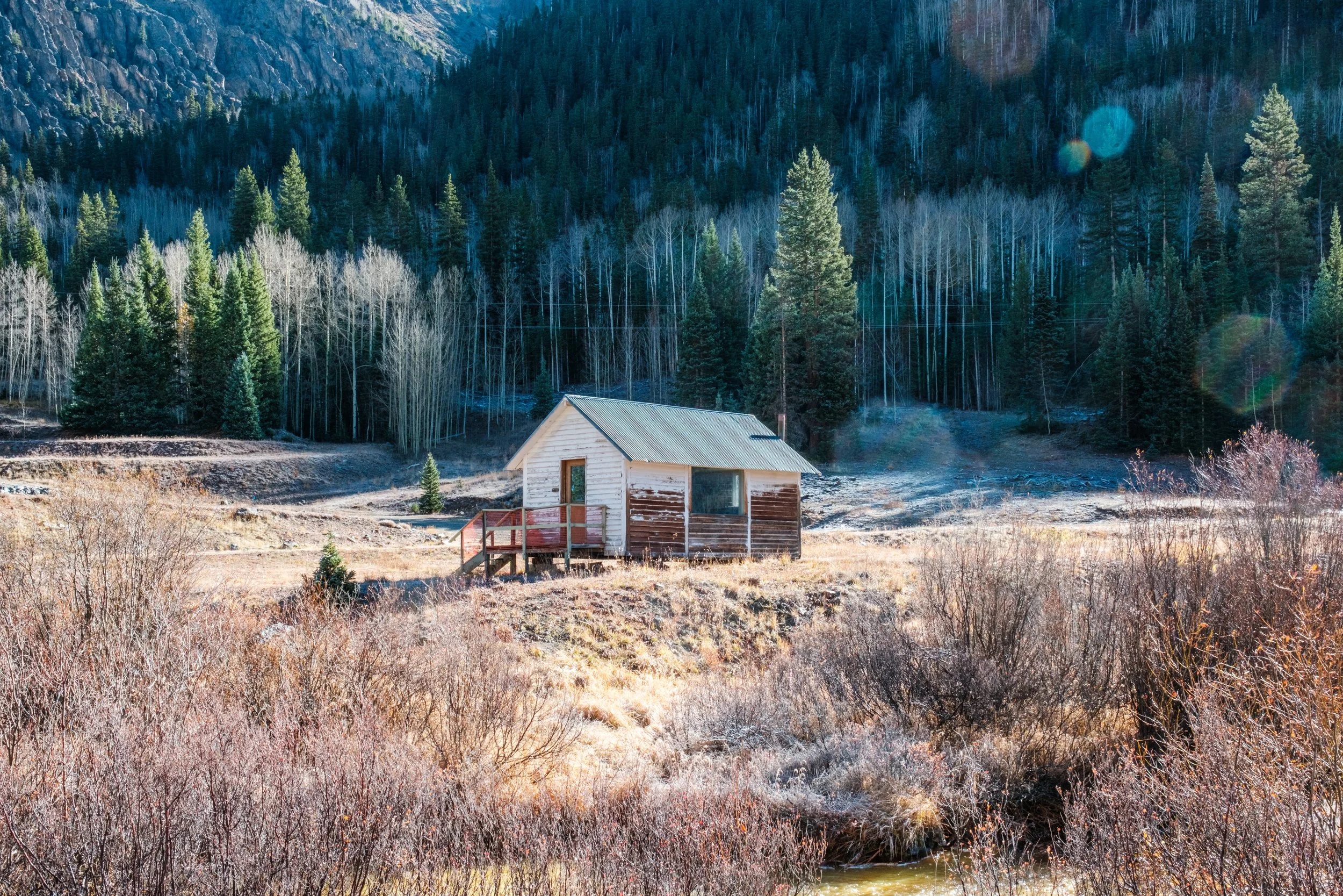 Small wooden cabin with a front porch, situated in a rural landscape with dry shrubs in the foreground and a forest of pine and deciduous trees on a mountainside in the background.