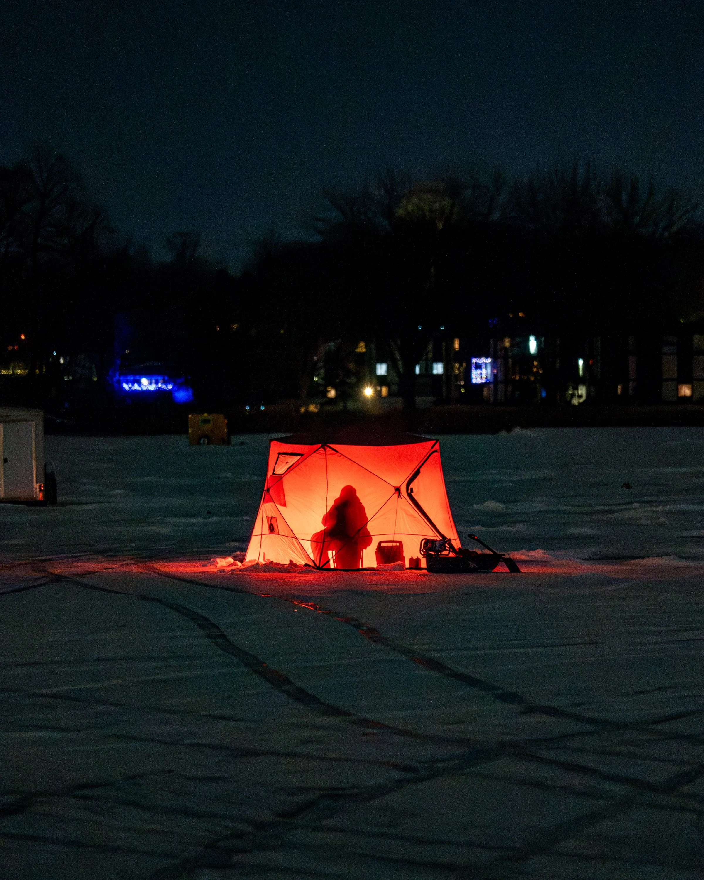 A person standing by a red illuminated tent on snow at night, with tire tracks in the snow and a dark background with trees and some distant lights.