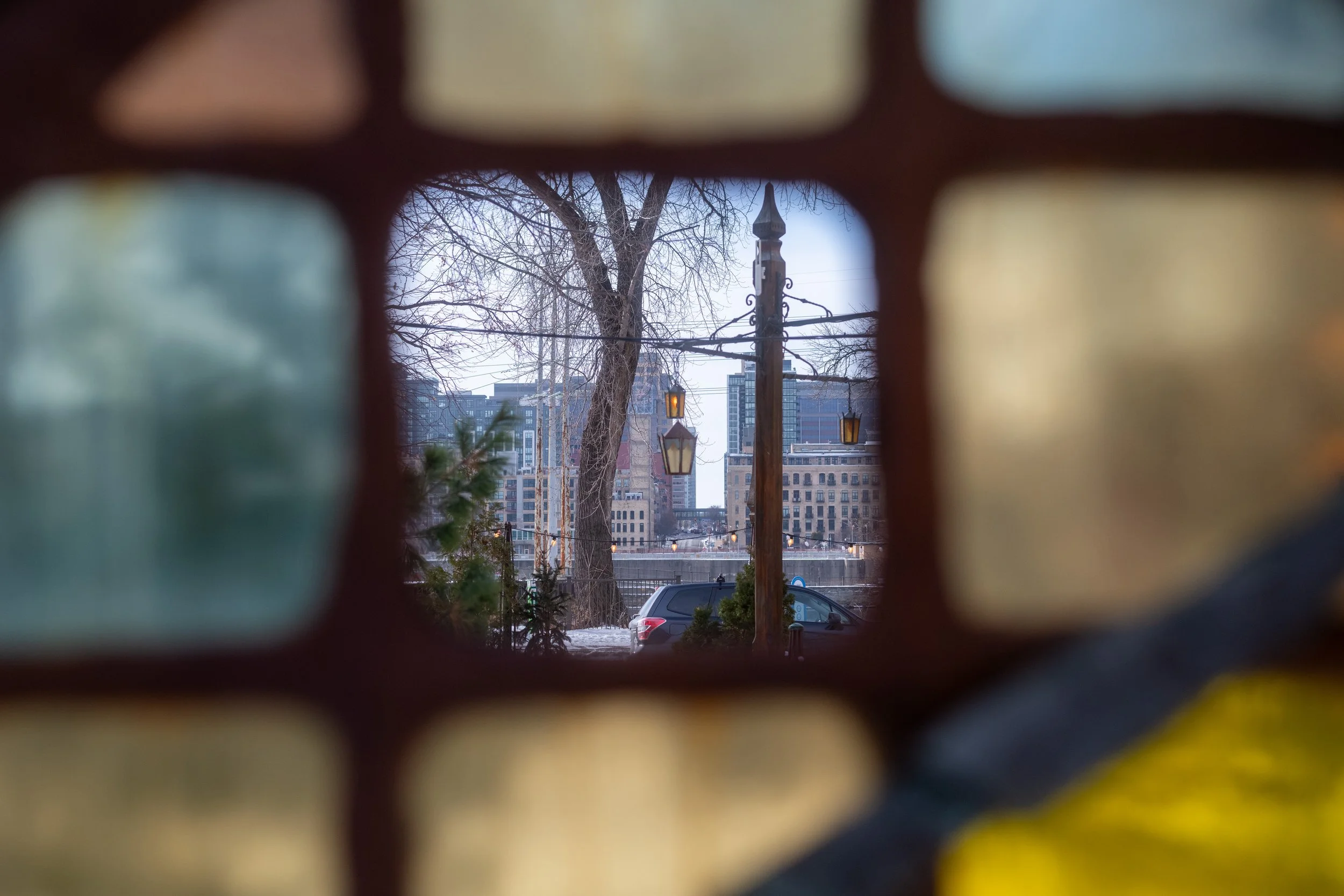 View through a small window showing a cityscape with tall buildings, leafless trees, street lights, and parked cars.