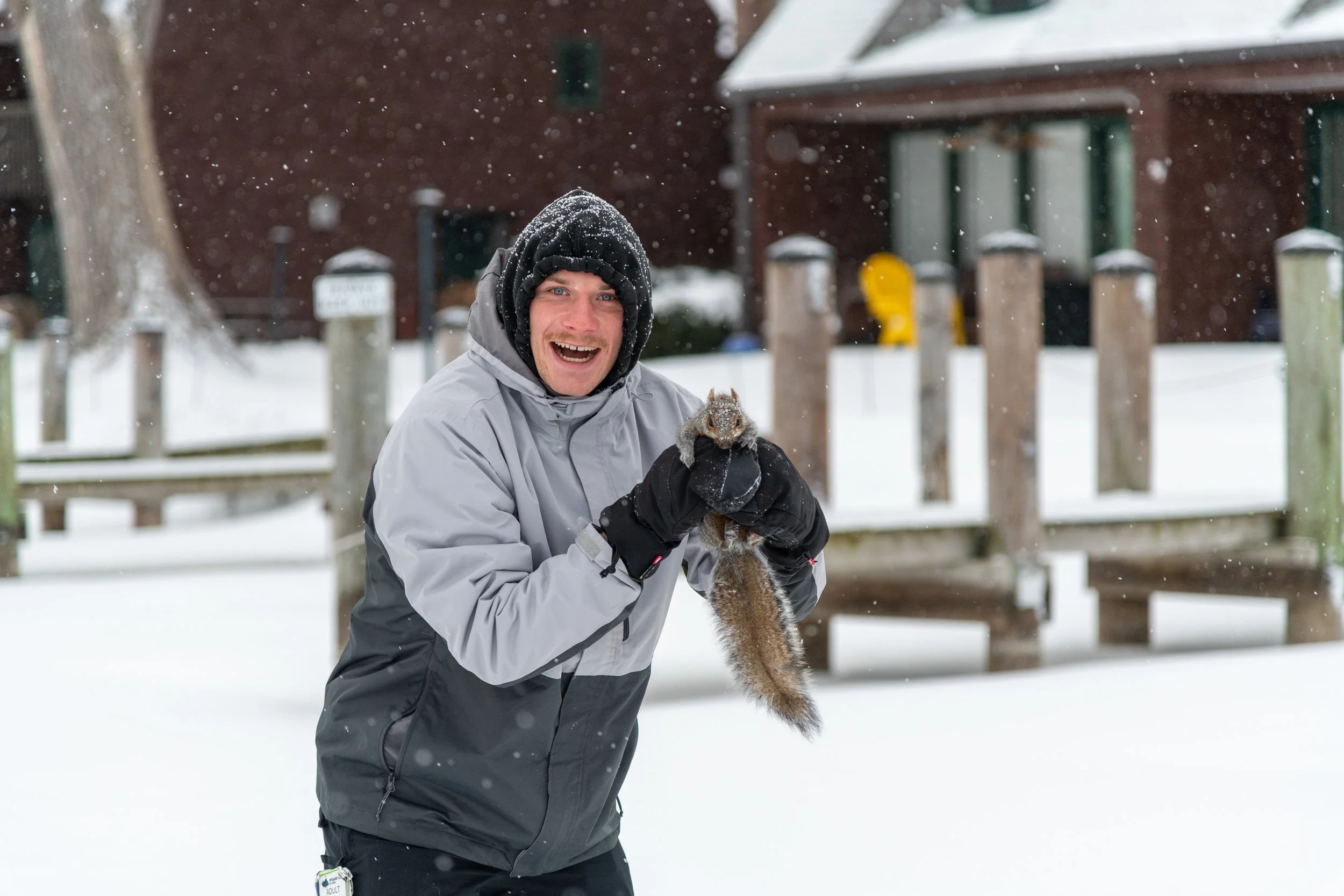 A man dressed in winter clothing standing in the snow, holding a squirrel with a joyful expression.