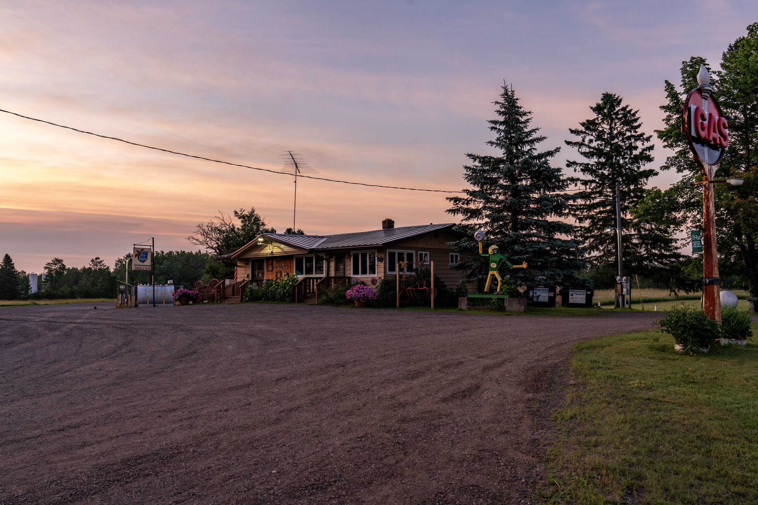 A small roadside establishment at sunset with a gravel parking area, a building with a porch, various decorations, a large sign reading 'GAS', and a colorful vintage-style figure with a green and yellow outfit near an electrical box.