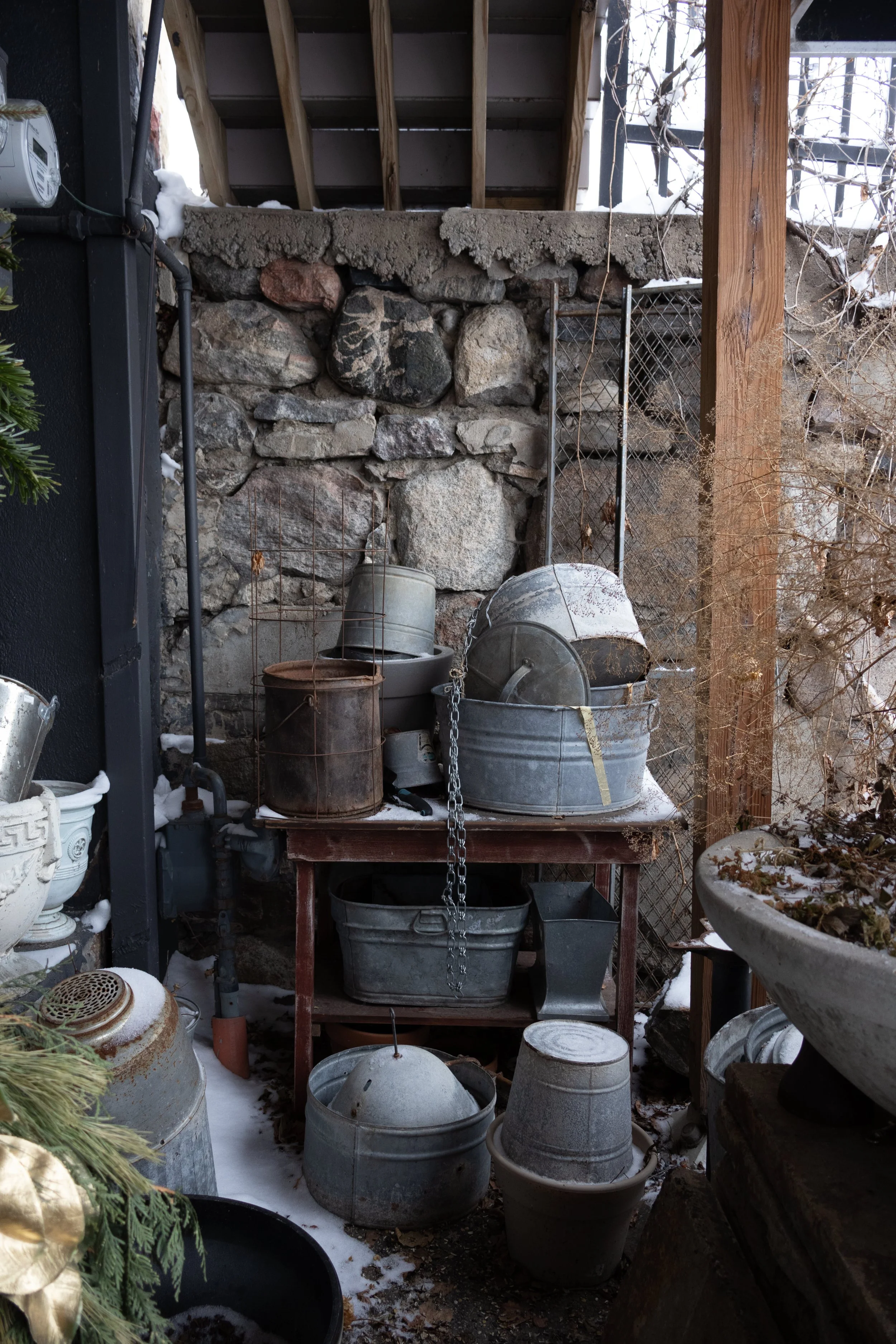 Outdoor garden storage area with metal buckets, tubs, and pots on a wooden table and the ground, surrounded by a stone wall, wooden posts, and wire fencing, with snow on the ground and some dried plants.