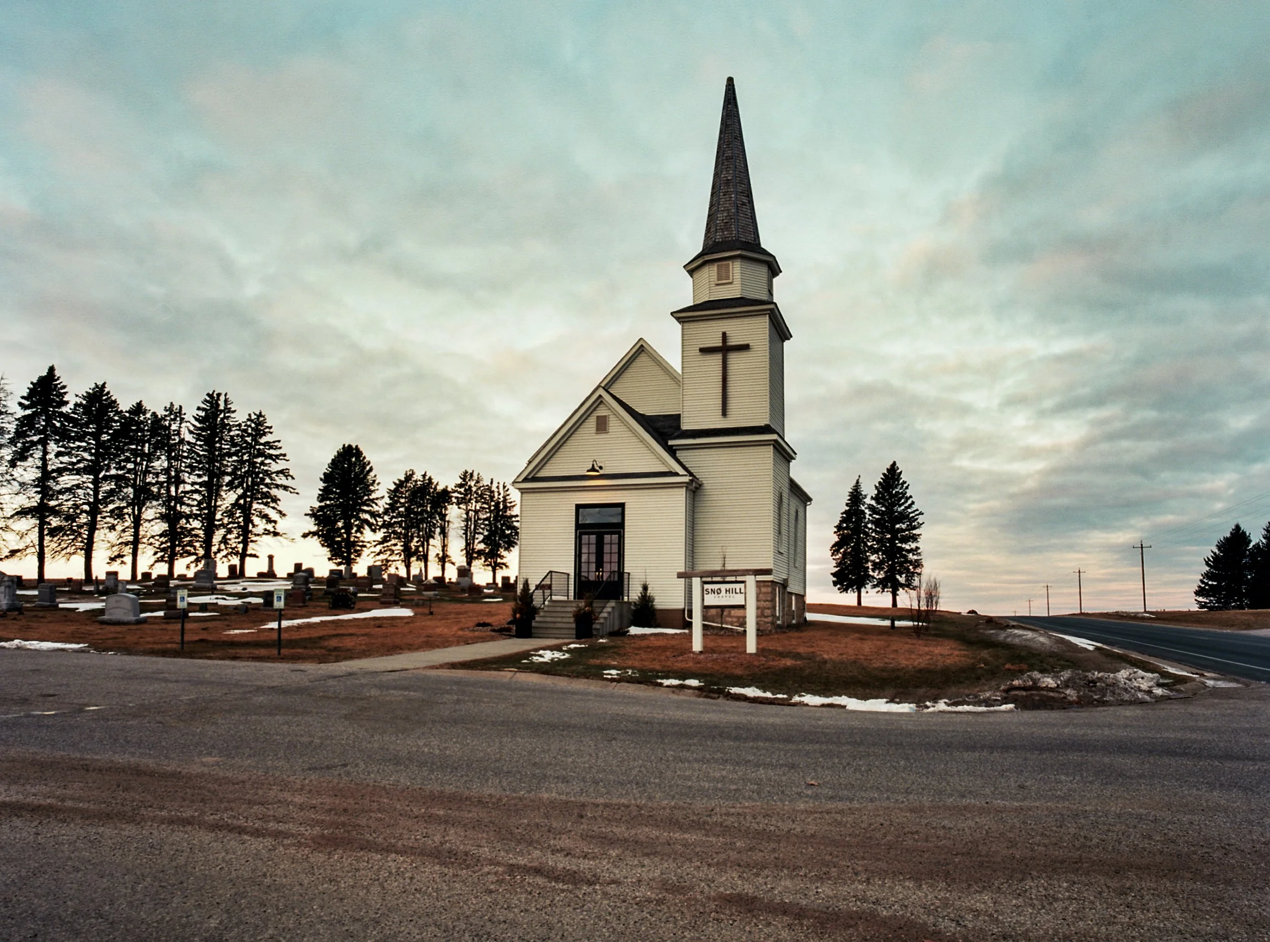 Small white church with a tall steeple, black cross, and steps leading to the door, situated on a hill with a sign reading 'Sno Hill'; graveyard with tombstones on the left, sparse trees, cloudy sky, and a road nearby.