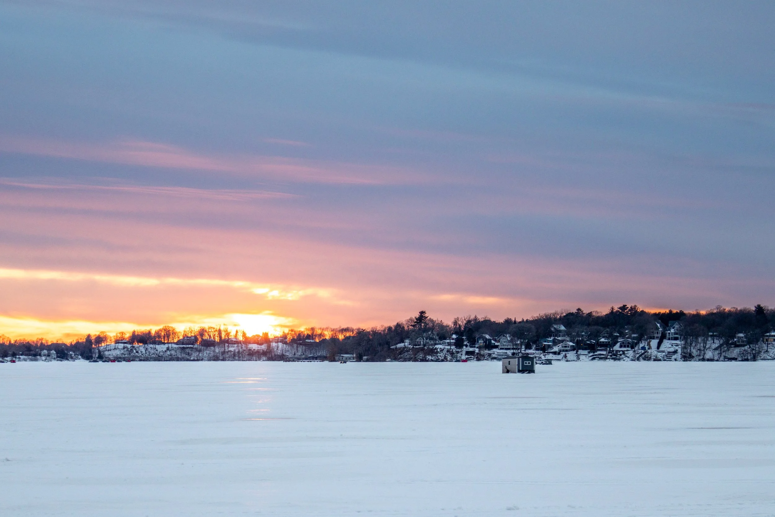 Sunset over a snow-covered frozen lake with a small house and houses on a hill in the background, with a colorful sky.