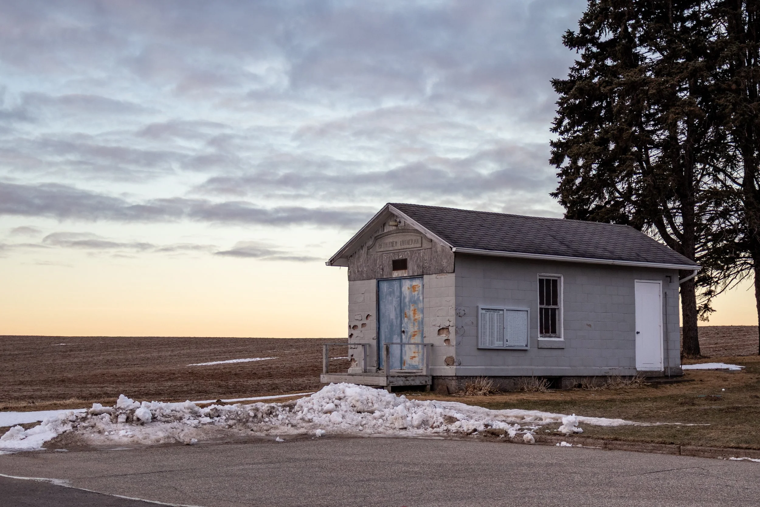 An abandoned small building with peeling paint on its door, situated in a rural area with snow on the ground and a large tree nearby, under a cloudy sky during sunset or sunrise.