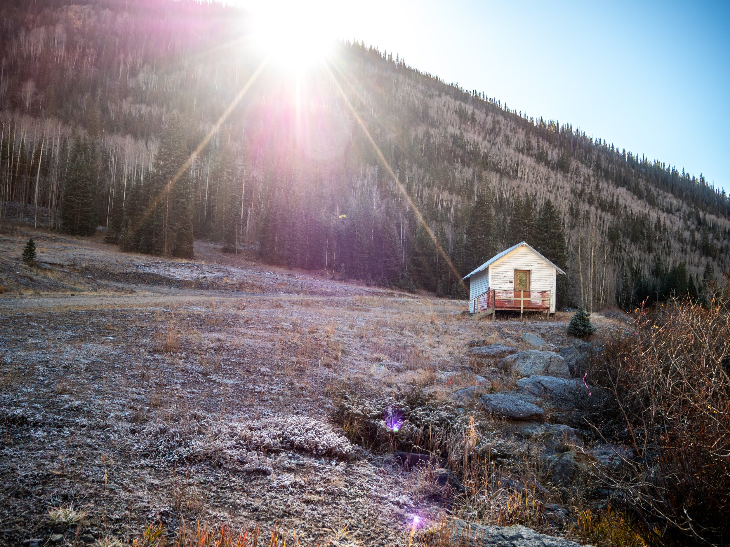 A small white wooden house with red railings on a hill, surrounded by rocks, dry grass, and sparse vegetation, with a forest of pine trees and a mountain in the background. The sun is shining brightly from above, creating lens flare effects.