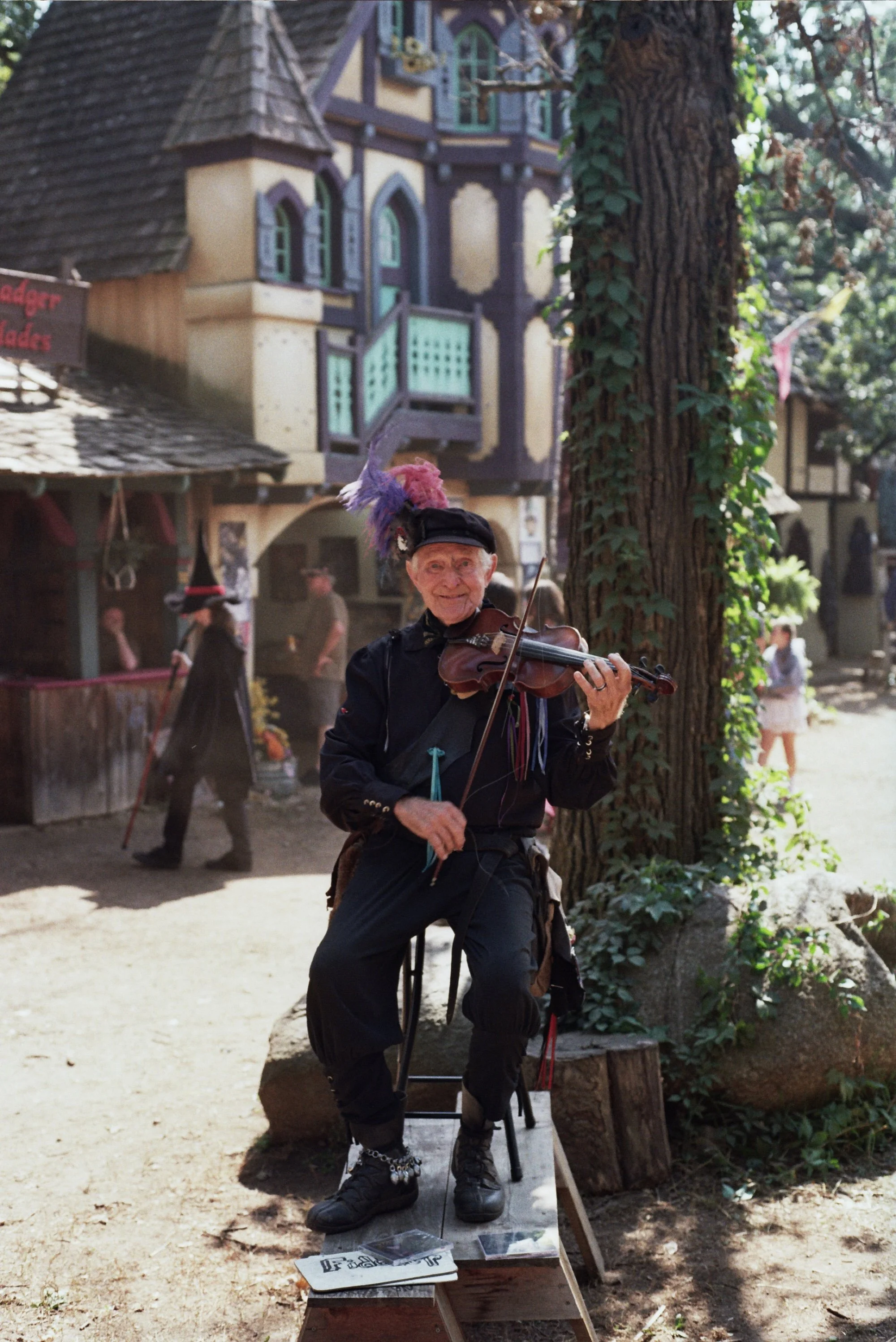 A street performer dressed in black, wearing a feathered hat, plays the violin on a small platform in a village-like setting with medieval-style buildings and people walking in the background.