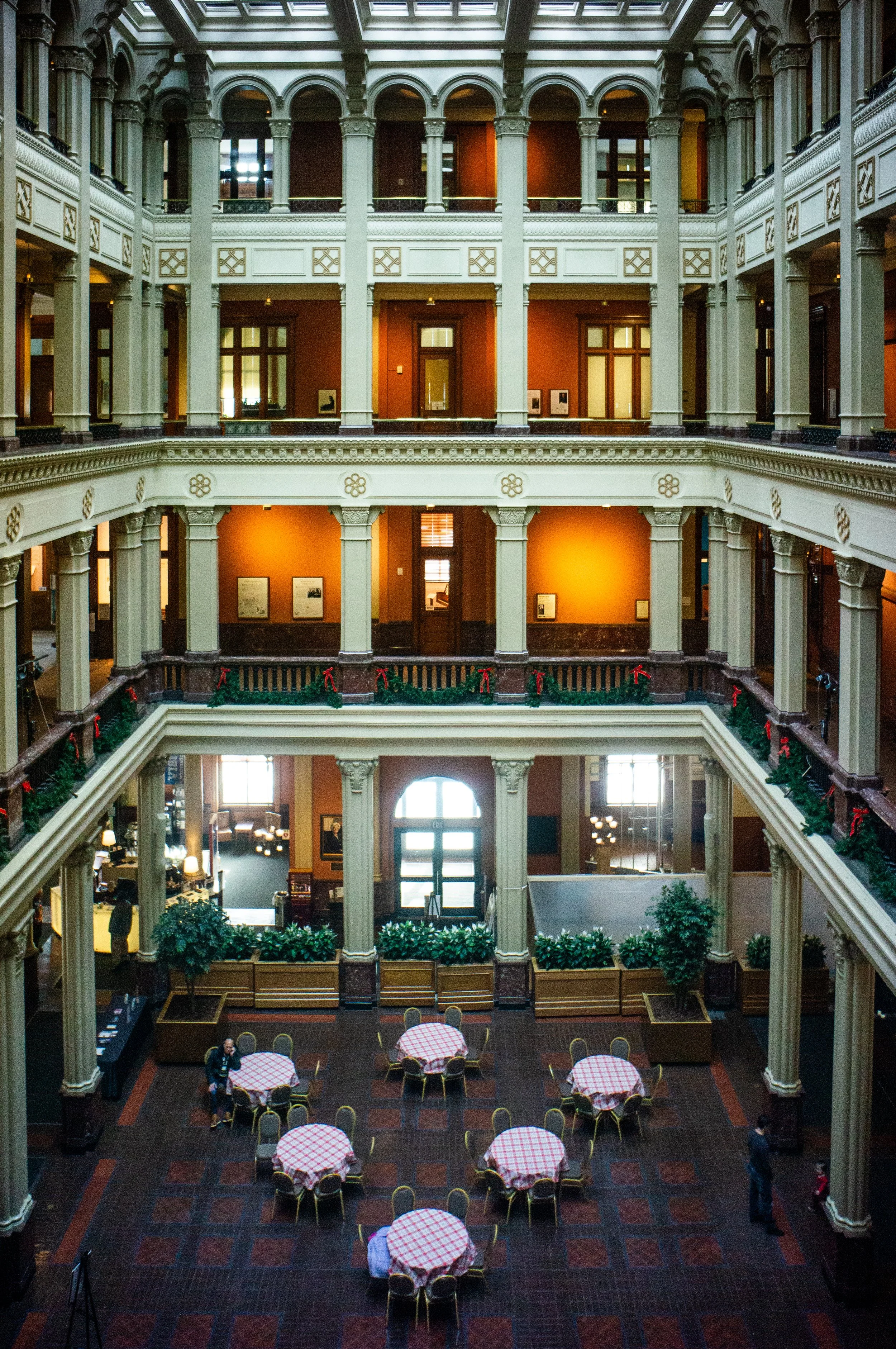 Interior of a historic building with multiple floors, ornate architecture, and large windows. The ground floor has round tables with checkered tablecloths and chairs, decorated for a festive event with Christmas garlands and ribbons along the railing