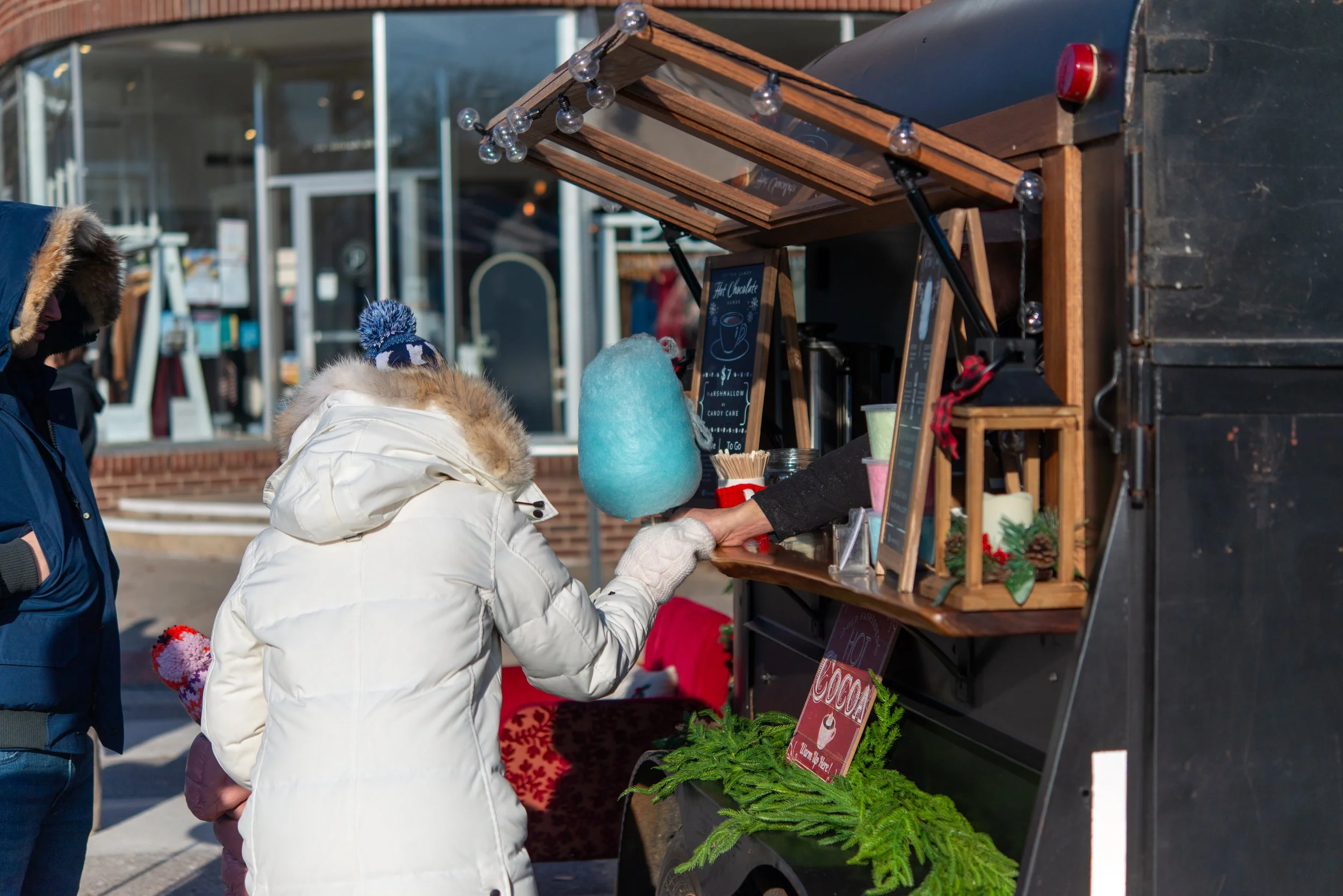 A woman in a white winter coat with a fur-lined hood and knit hat is buying cotton candy from a street vendor cart. The vendor's hand is visible, handing over the cotton candy. The cart is decorated with holiday greenery and signs. Two people are wai
