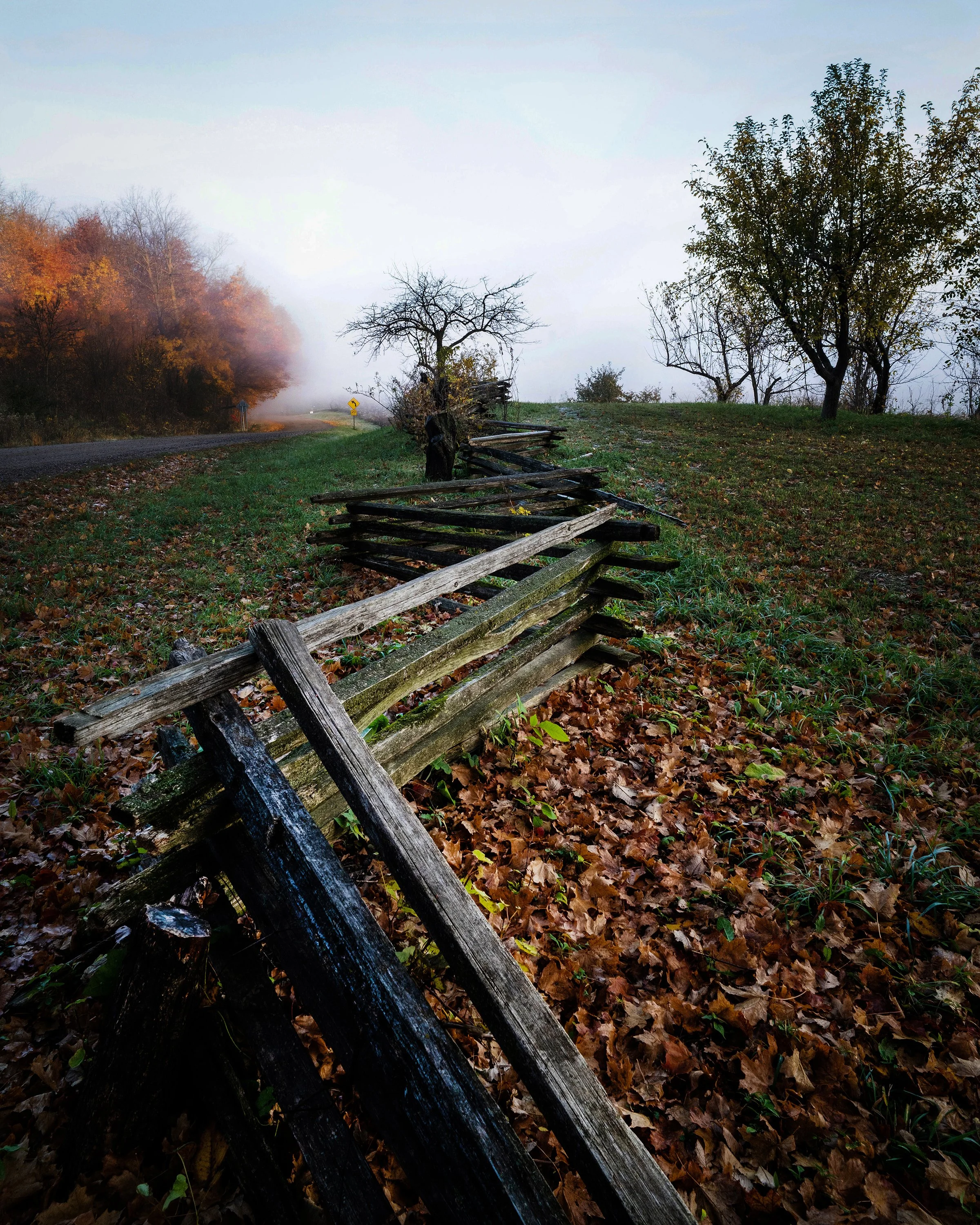 A fallen wooden fence along a rural roadside with autumn trees and fog in the distance.
