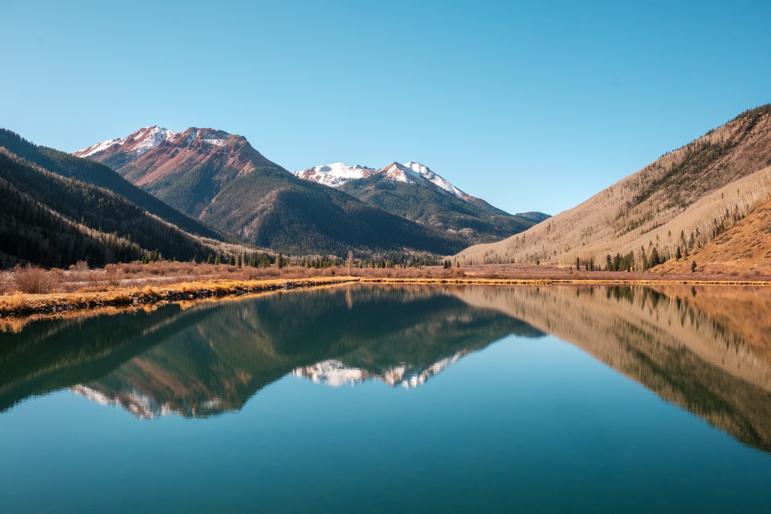 A calm lake reflecting mountains with snow caps under a clear blue sky.