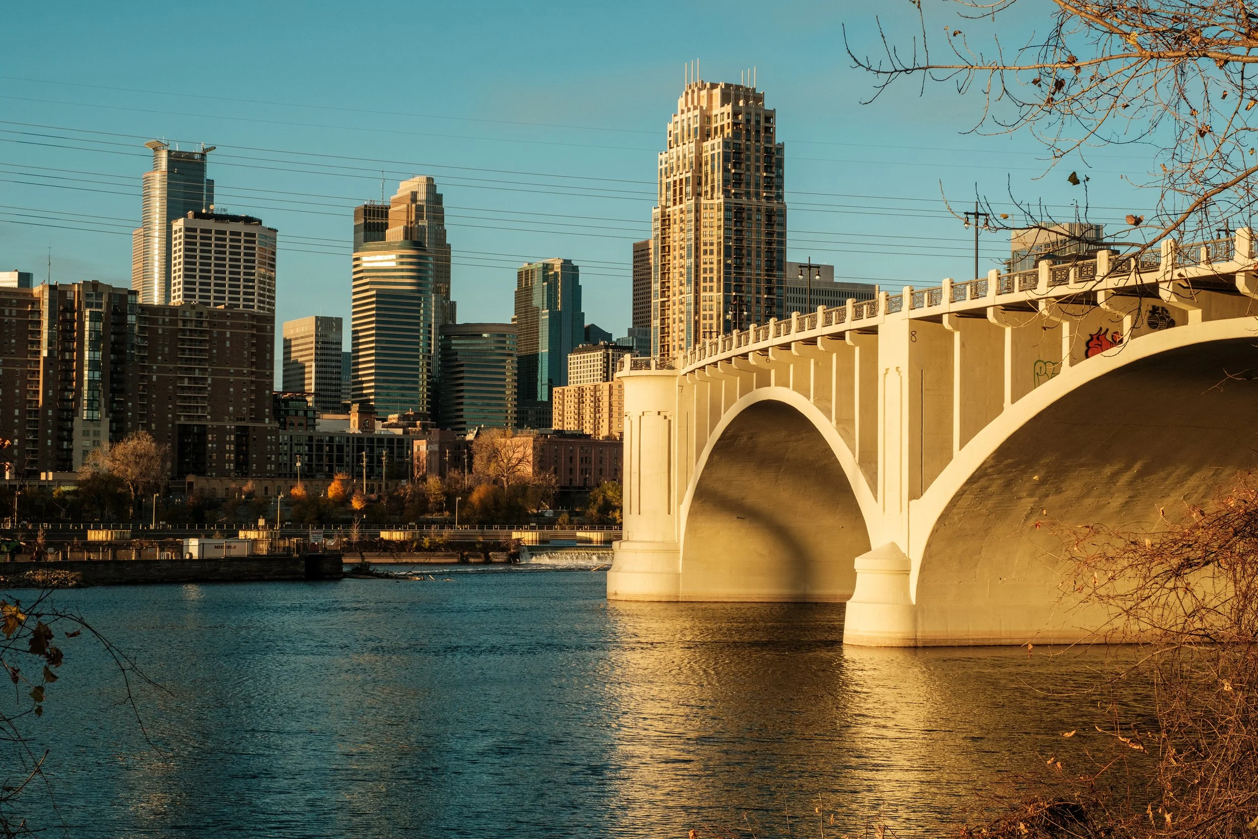 A city skyline featuring tall skyscrapers and a bridge over a body of water, with some trees and power lines in the foreground.